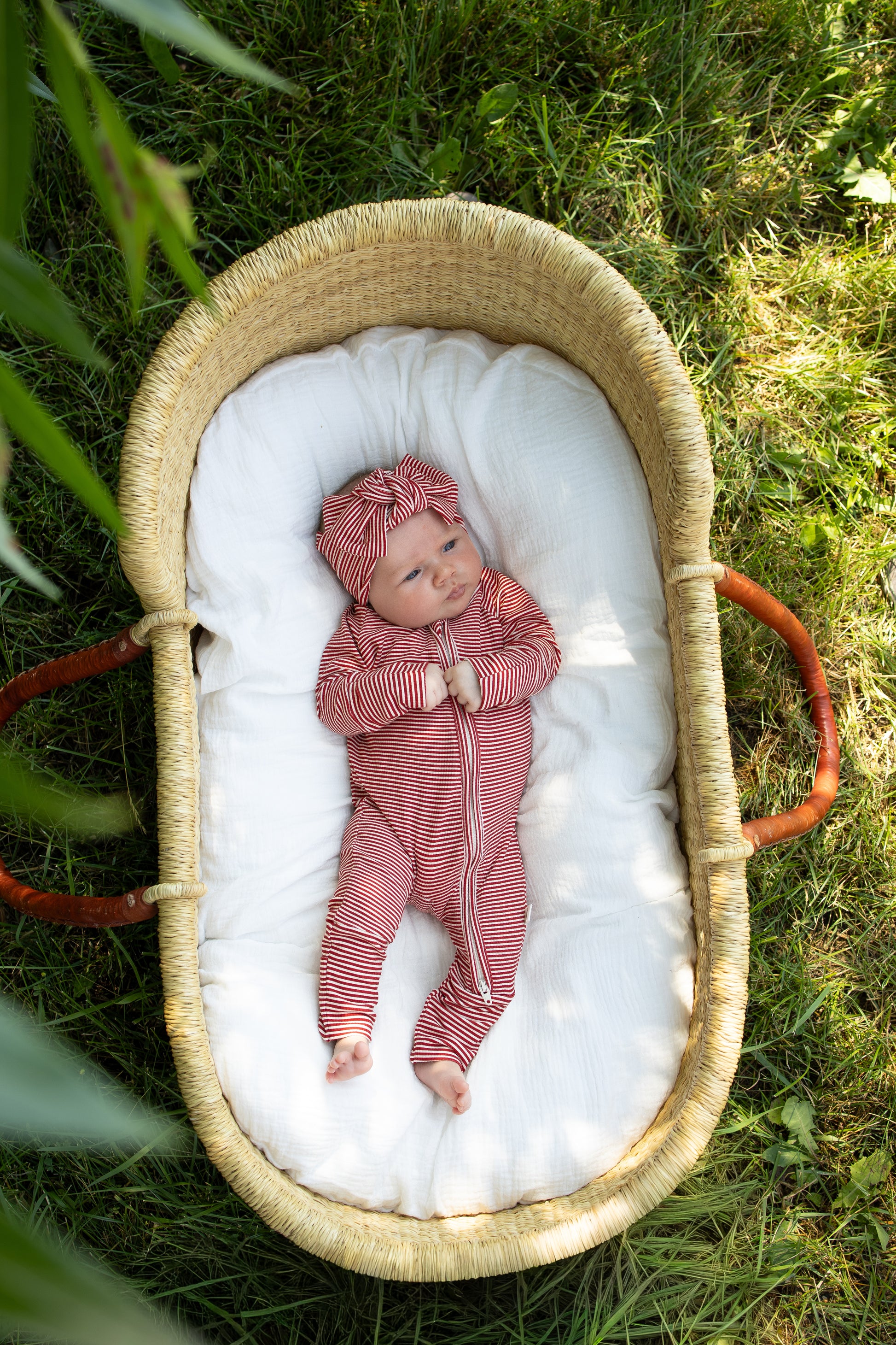 A baby wearing the forever french baby Bamboo Zip Pajamas in Peppermint Stripe Ribbed, along with a matching headband, rests in a woven basket lined with white bedding on green grass outdoors in daylight.