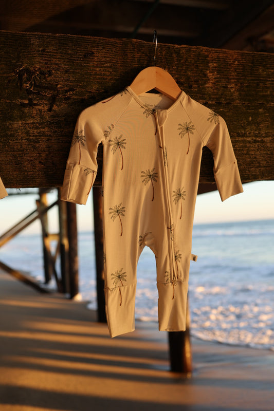 The Bamboo Zip Pajamas | Palm Tree by forever french baby, a beige hypoallergenic baby onesie with a palm tree pattern, hangs on a wooden hanger under a pier with sunlight and the ocean in the background.