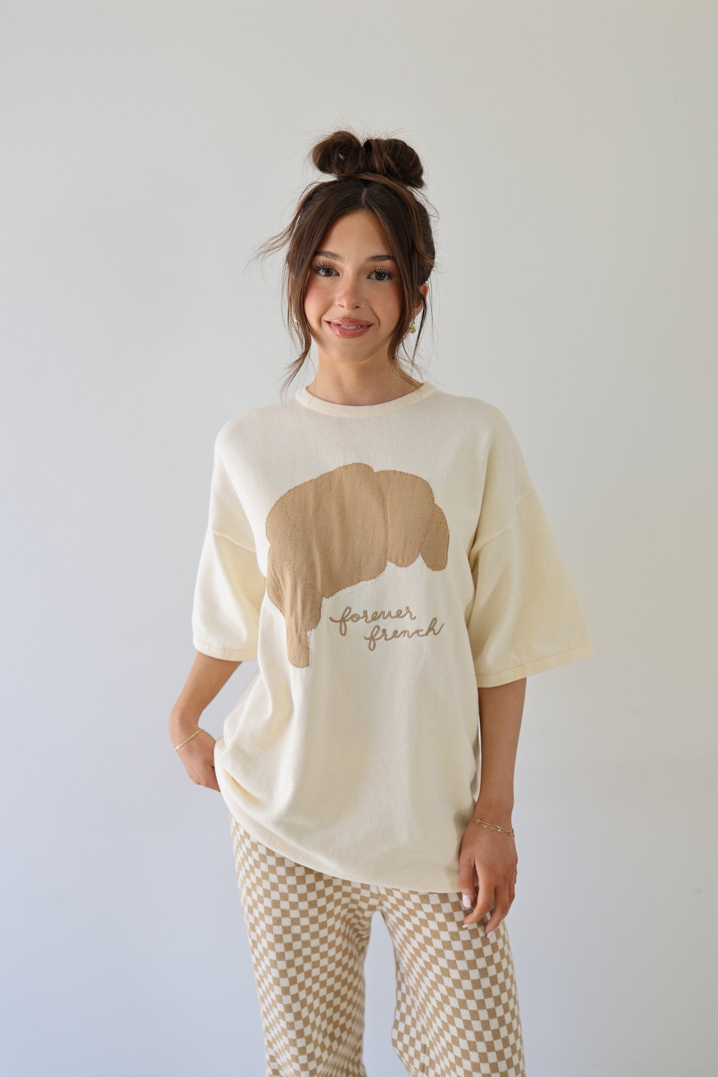 A young woman with long brown hair in a messy bun wears the Women's Knit Pant Set | Croissants by forever french baby, featuring an oversized cream top with a bear graphic and "forever friend" paired with beige checkered pants, smiling against a white background.
