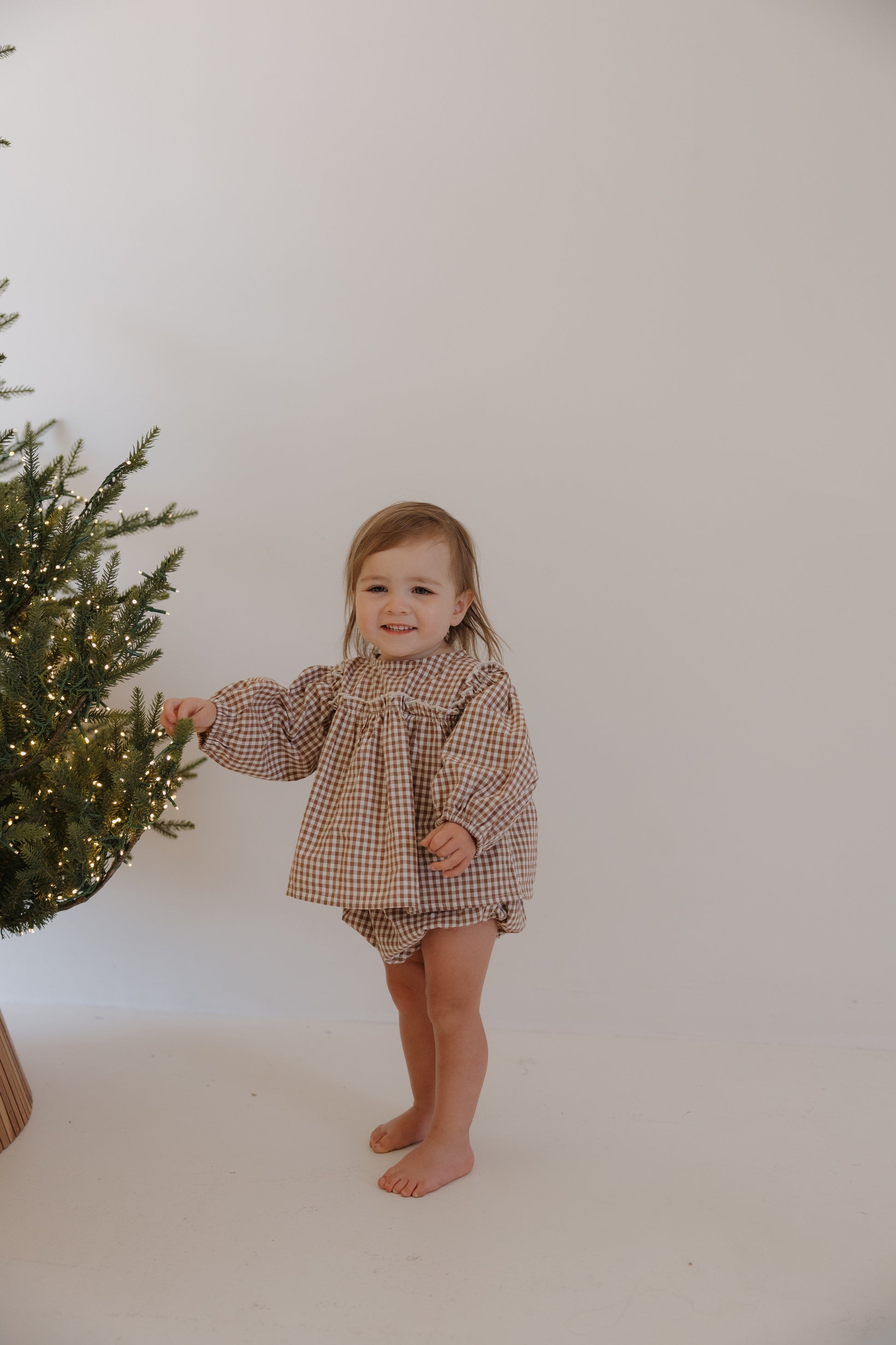 A smiling toddler in the forever french baby Bloomer Set | Honey Gingham stands barefoot beside a decorated Christmas tree with string lights, touching a branch in a bright room with a plain white wall.