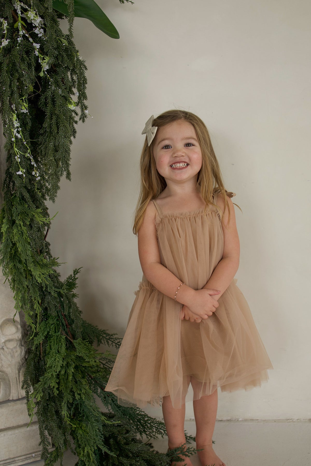 A smiling young girl wearing a beige forever french baby Teddy tutu dress with bow straps, standing beside a pine garland.