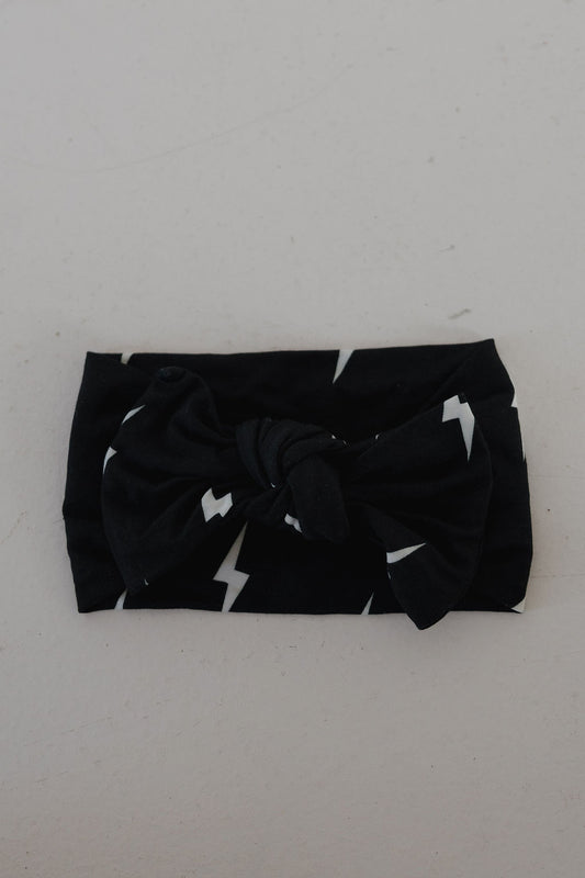 The Bamboo Head Wrap | Midnight Bolt by forever french baby features a black fabric with white lightning bolts and a large knotted bow, displayed on a light surface.