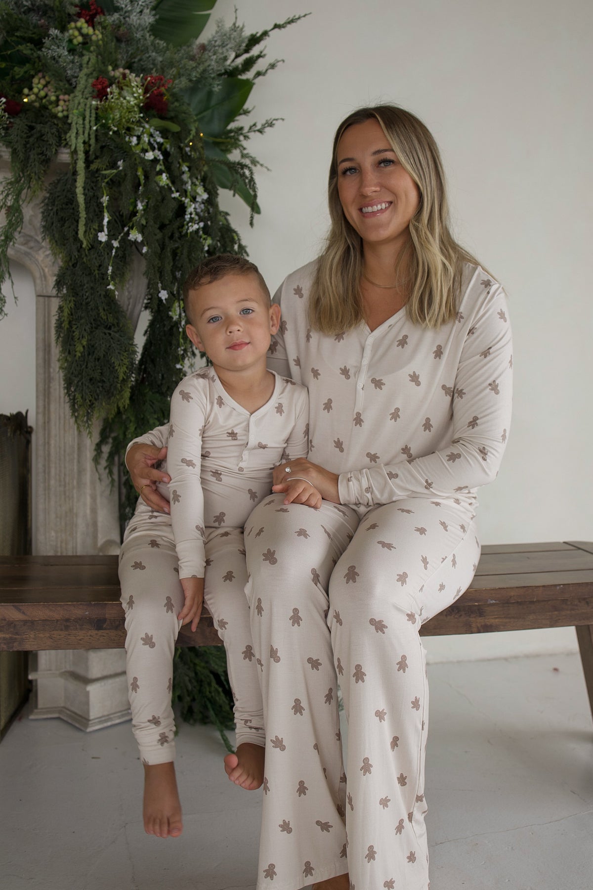 A woman and a young boy sit together on a bench wearing Bamboo Two Piece Pajamas | Gingersnap, patterned with small gingerbread men.