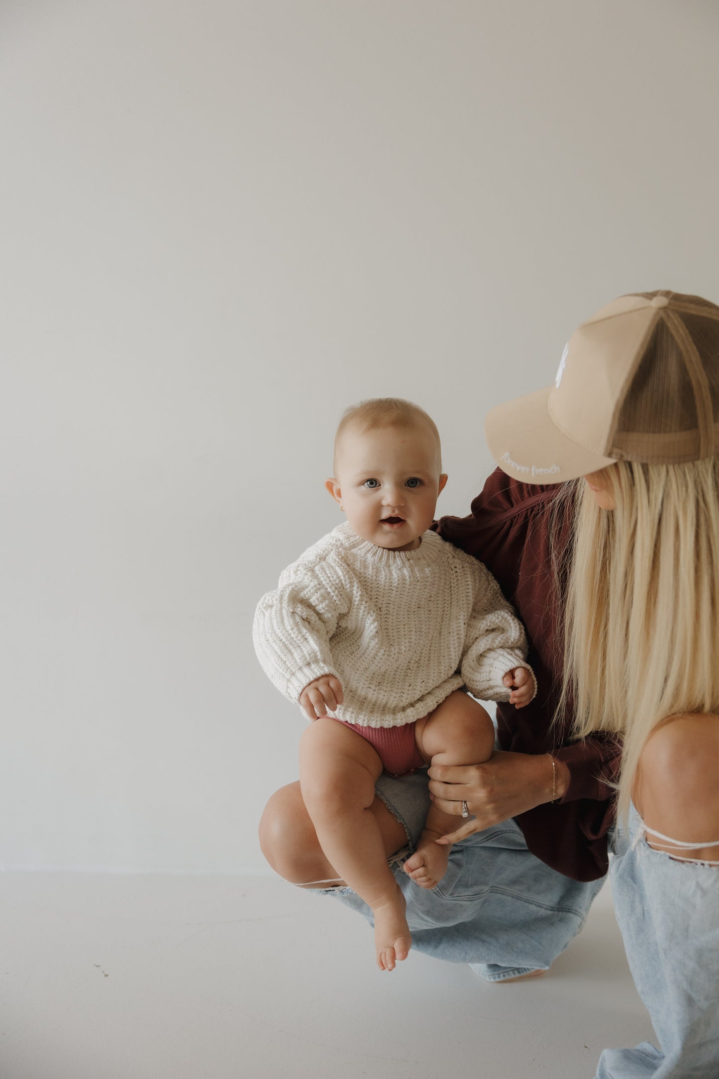 A baby in the forever french baby Child Knit Sweater | Cloud Confetti sits on a woman's knee. The woman, with long blonde hair and a beige cap, looks at the baby in a minimal, light-colored room.