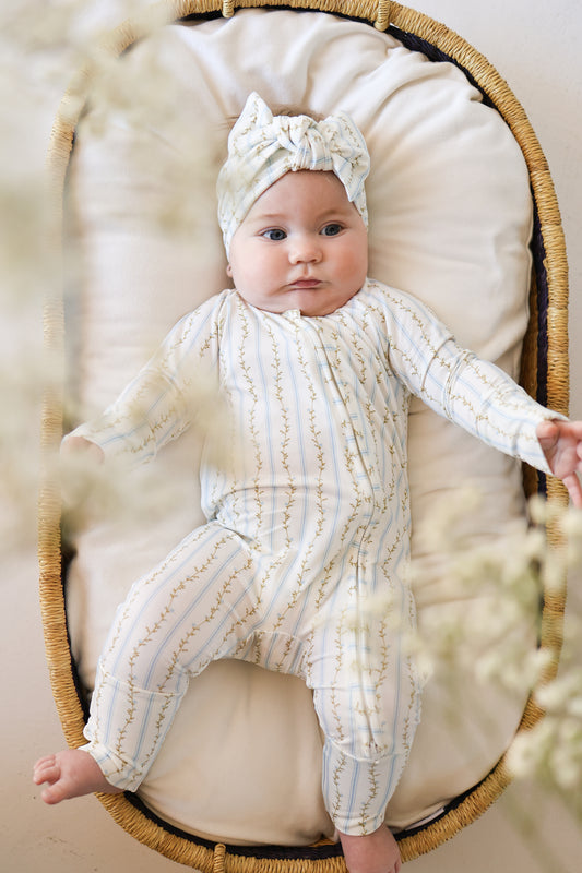 A baby lies calmly in a cushioned basket, wearing forever french baby’s Bamboo Zip Pajamas | Dainty Vines—breathable sleepwear with a white and yellow vine pattern—as soft flowers frame the scene.