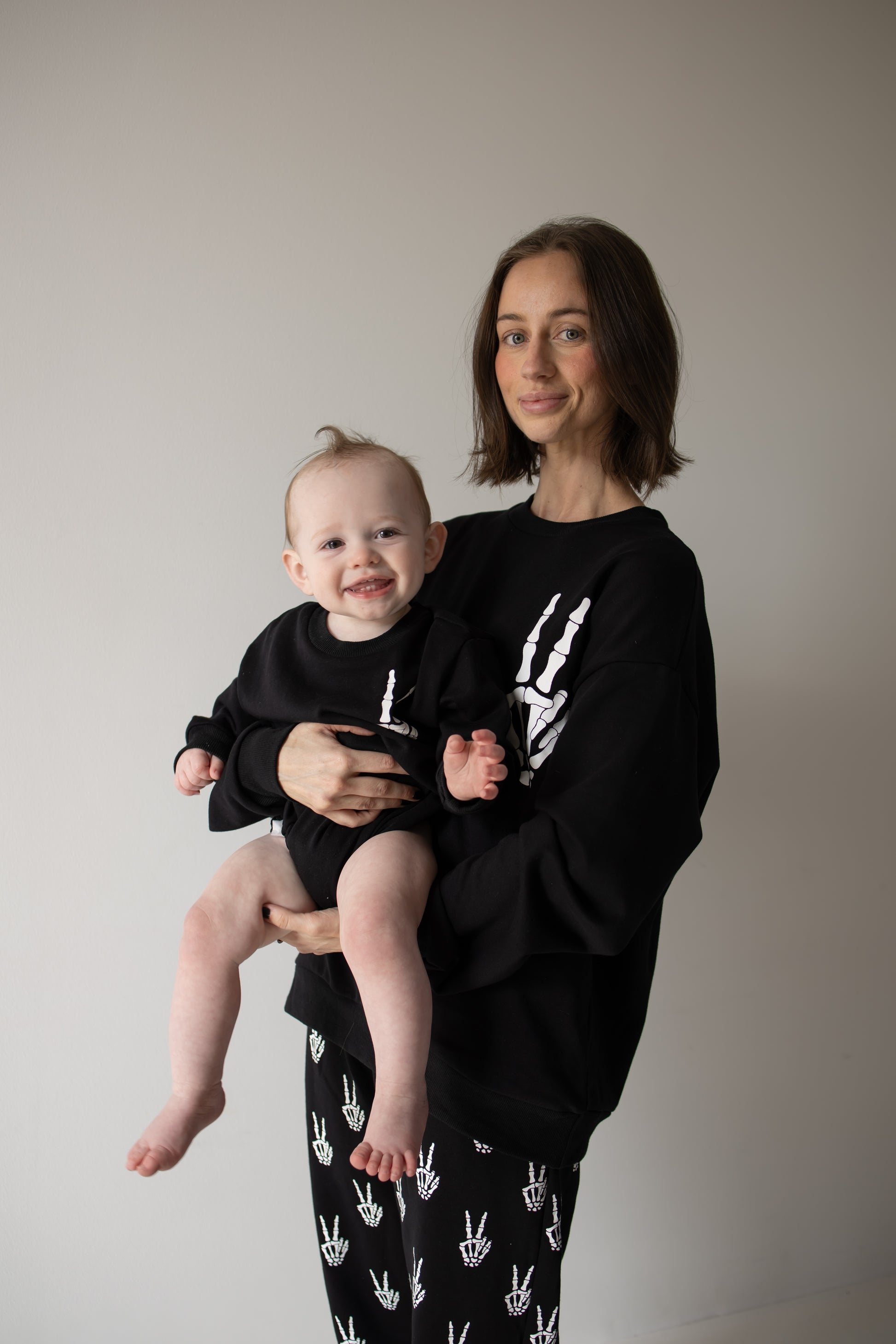 A woman holds a smiling baby, both dressed in matching “Midnight Skelly” sweatshirt rompers from forever french baby, featuring playful white handprint designs on soft black cotton. They stand against a plain, light background.