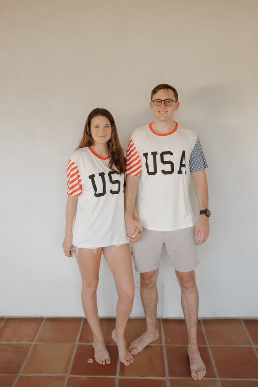 A woman and man stand barefoot on a tiled floor, holding hands, both wearing matching forever french baby Adult Bamboo T-Shirts | Stars & Stripes with red, white, and blue details.