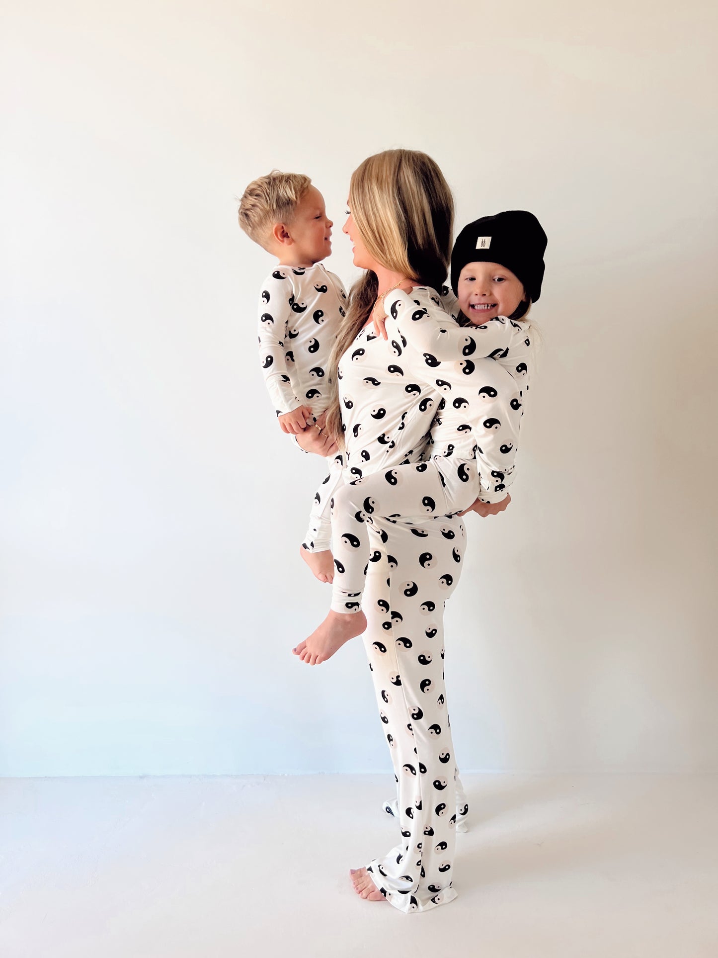 A woman stands in profile carrying two young children, all wearing matching Forever French Baby Women's Bamboo Pajamas in black and white yin-yang print. The child on her left is smiling and wearing a black hat. The background is a plain, light-colored wall.