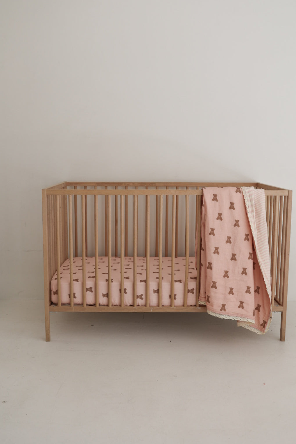 A wooden baby crib with vertical slats features the forever french baby Muslin Quilt | Pink Lottie & Sadie Stripe, plus a matching pink sheet. The quilt’s subtle stripes add charm to your nursery against a simple, light wall.