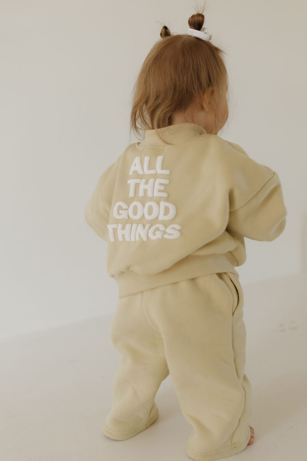 A toddler in a beige "Child Sweat Set | All the Good Things" by forever french baby, featuring the phrase "ALL THE GOOD THINGS" printed on the back. The child, with two small ponytails, stands on a light-colored floor, facing away from the camera.