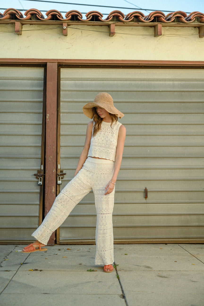 A person wearing a straw hat, a sleeveless crochet button back top from MIOU MUSE, and matching 100% cotton pants stands in front of a closed garage door. They lean slightly with one leg crossed in front. The setting is sunny, and they wear orange sandals.