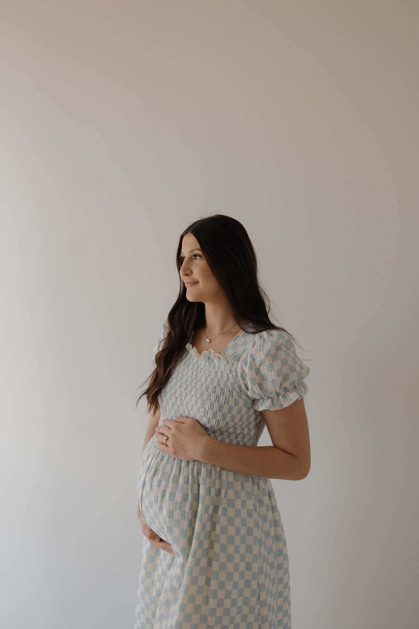 A pregnant woman with long brown hair wears the forever french baby Women's Short Sleeve Muslin Dress in Morning Check, standing against a plain light background as she gently holds her belly and gazes serenely to the side.