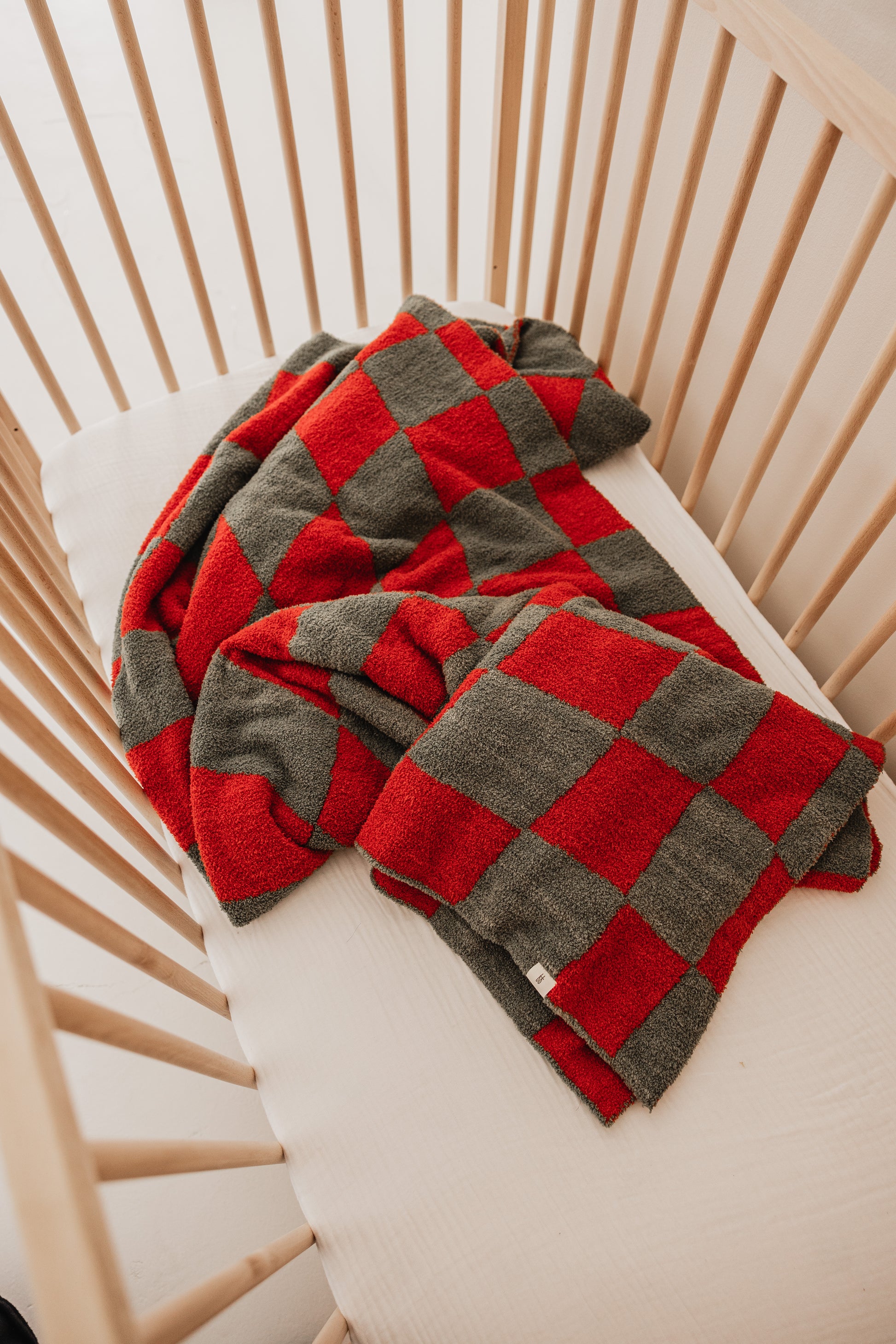 In a wooden crib with a white mattress, the cozy Retro Christmas | Plush Blanket from forever french baby is neatly draped over the side. Made with microfiber feather yarn, this OEKO Standard 100 certified piece adds a touch of festivity and complements the crib's simple design with its red-and-gray checkered pattern.