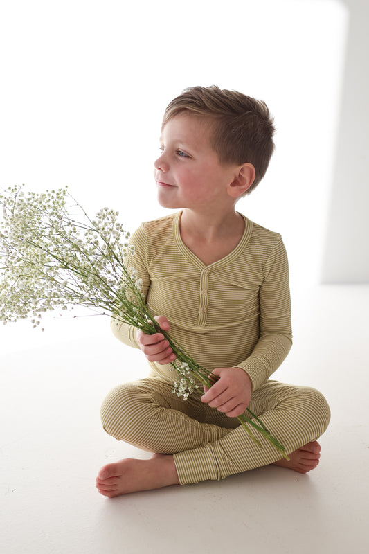 A young child in the Willow Stripe Ribbed Bamboo Two Piece Pajamas by forever french baby sits cross-legged on a white floor, holding a bouquet of small white flowers and smiling while looking to the side.