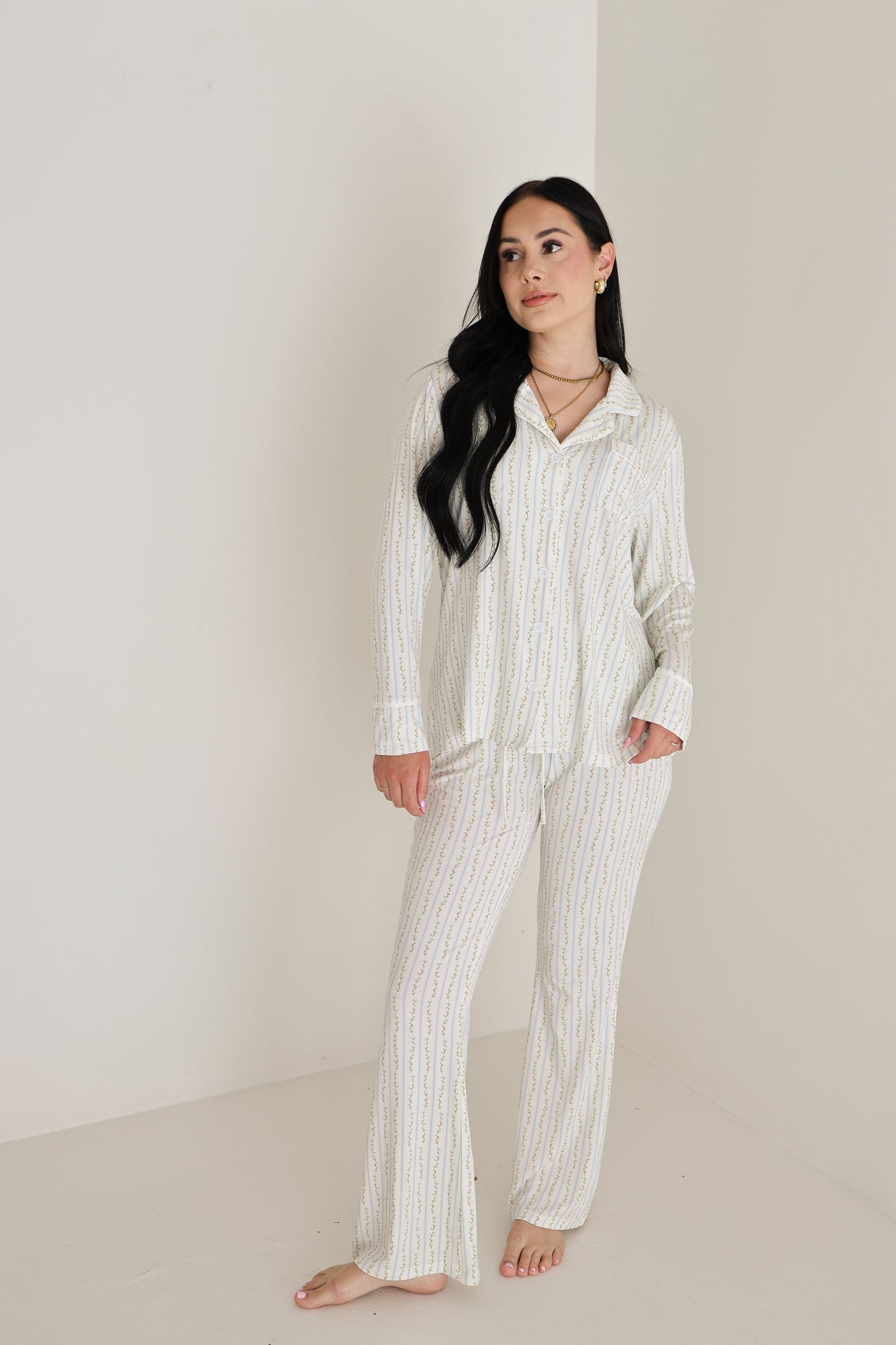 A woman with long dark hair stands barefoot in a minimal, light room, wearing the forever french baby Women's Bamboo Button Up Lounge Set | Dainty Vines. She looks slightly to the side with a neutral expression.