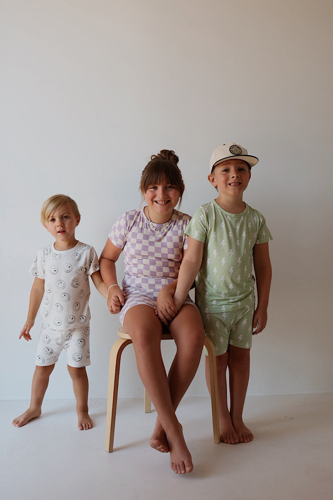Three young children pose indoors against a plain background. The child on the right wears the forever french baby Bamboo Short Set in Grape Soda, while the others sport playful, breathable sleepwear with smiley faces and purple checks.