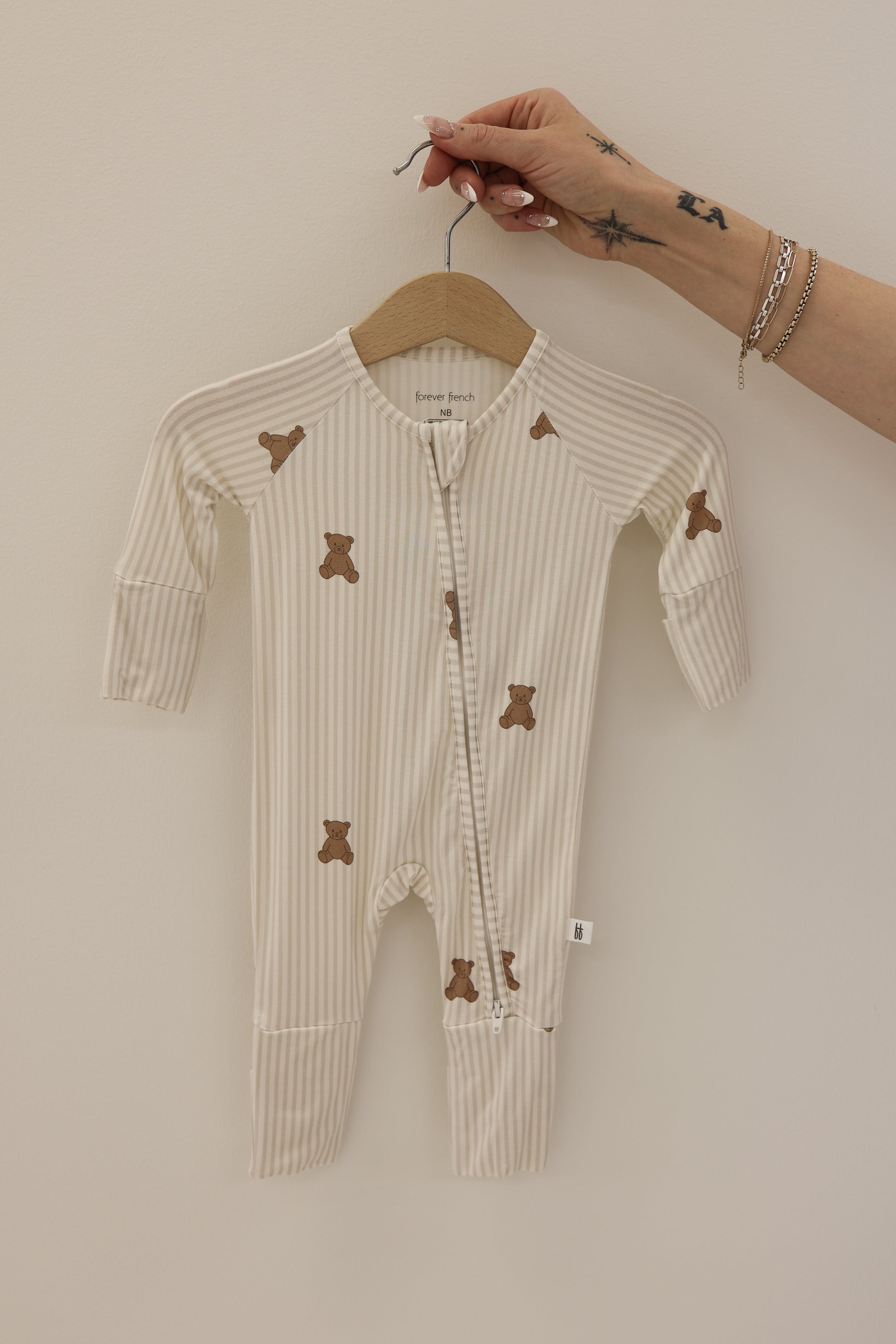 A hand holds forever french baby Bamboo Zip Pajamas | Teddy—a beige and white striped footed onesie featuring small brown teddy bear prints—on a wooden hanger, displayed against a plain light background.