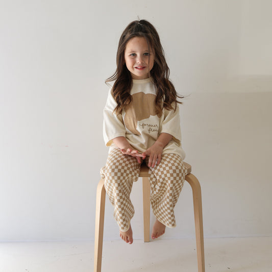 A young girl with long brown hair sits barefoot on a wooden stool against a white background, smiling in the forever french baby Knit Pant Set | Croissants, featuring an oversized tan graphic top and checkered beige-and-white pants.