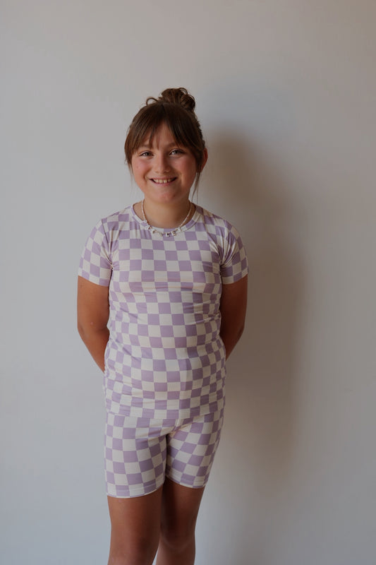 A young girl with light brown hair in a bun smiles at the camera, wearing the forever french baby Bamboo Short Set in Grape Soda—a short-sleeved, breathable lavender and white checkerboard sleepwear—against a plain light wall.