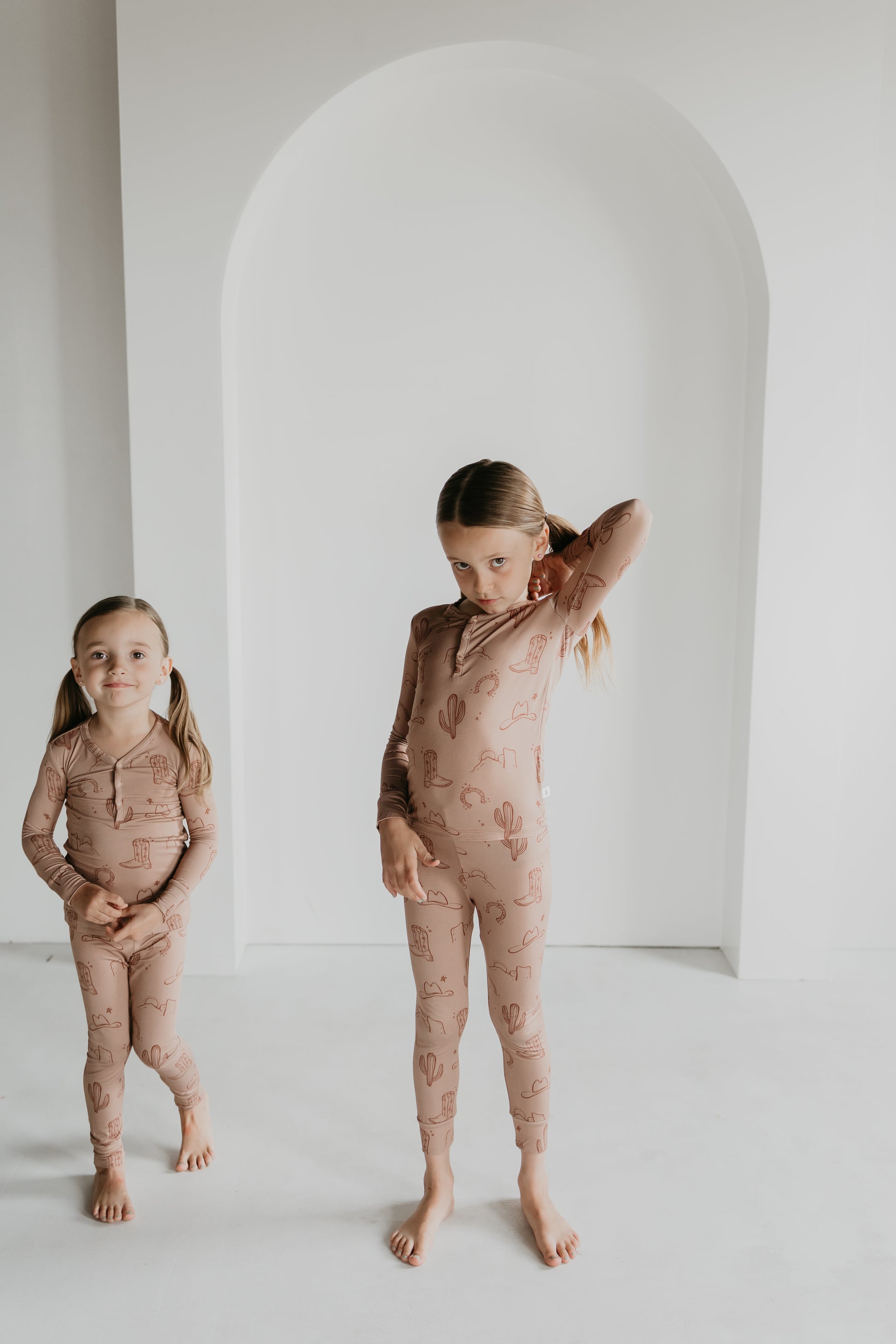 Two young children stand indoors in a minimally decorated room with a white arched doorway in the background. Both wear matching beige Bamboo Two Piece Pajamas from Minty x FF Wild West Desert Dreams, adorned with a cactus pattern. The child on the right has one hand on their head, while the other stands with hands by their sides.