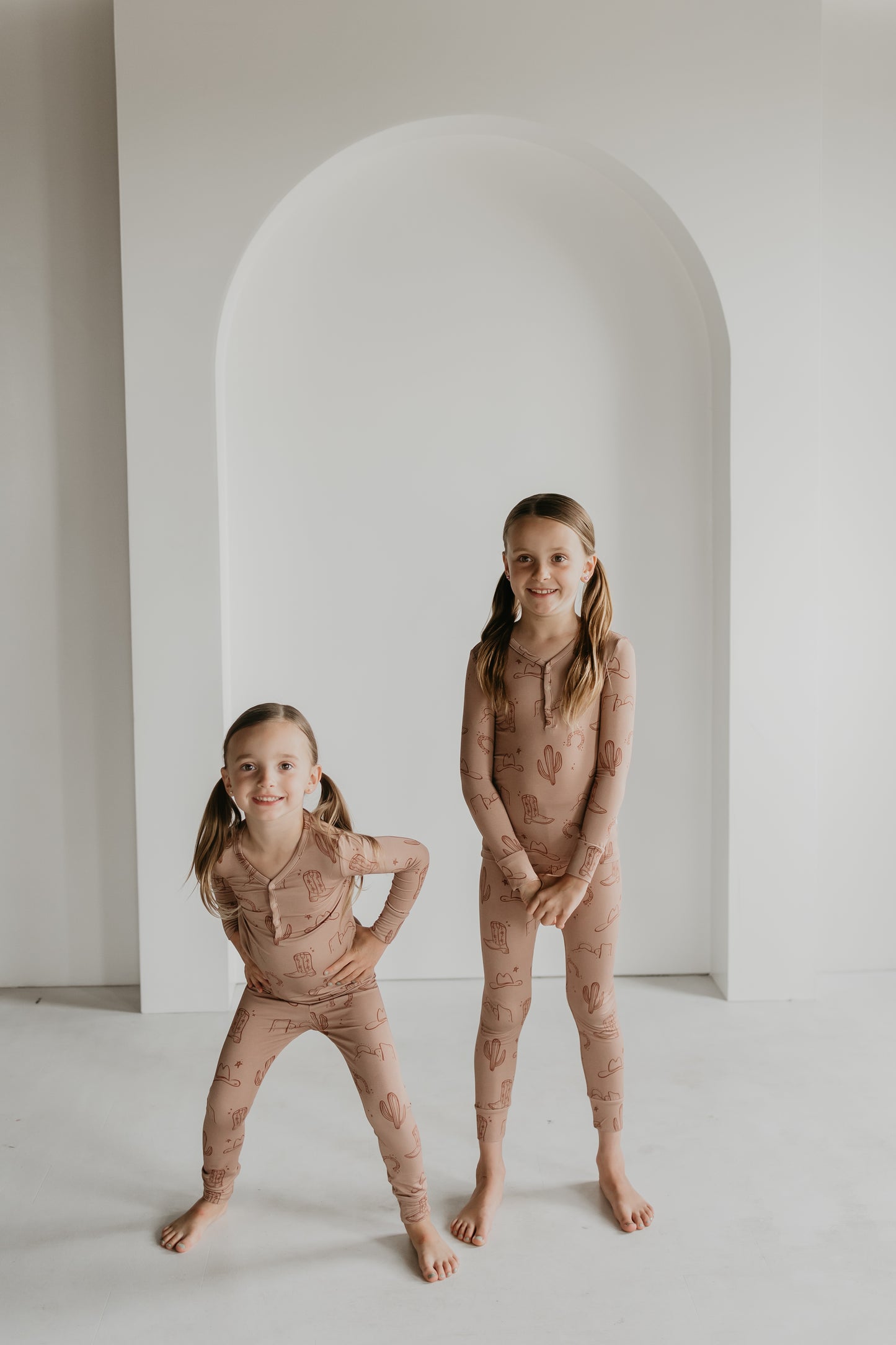 Two young girls stand in a minimalist room with a white arch backdrop. Both are dressed in matching beige bamboo pajamas adorned with a cactus pattern from the Minty x FF Wild West Desert Dreams collection. The younger girl on the left, sporting pigtails, leans forward playfully while the older girl stands tall and smiles.