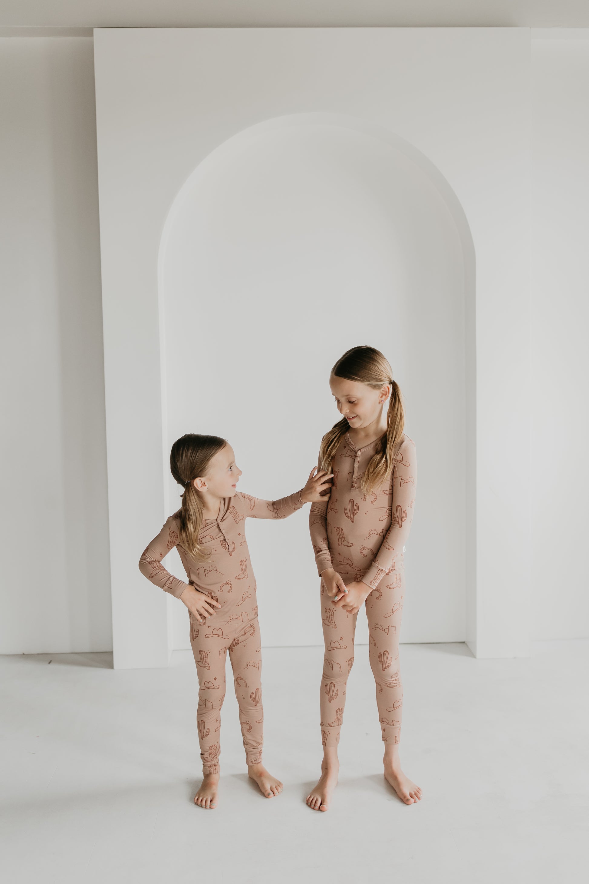 Two young girls in matching Bamboo Two Piece Pajamas from the Minty x FF Wild West Desert Dreams collection, decorated with pink cactus patterns, stand barefoot in front of a white archway. The older girl, with pigtails, smiles affectionately as the younger girl, also with pigtails, touches her shoulder while looking up at her.