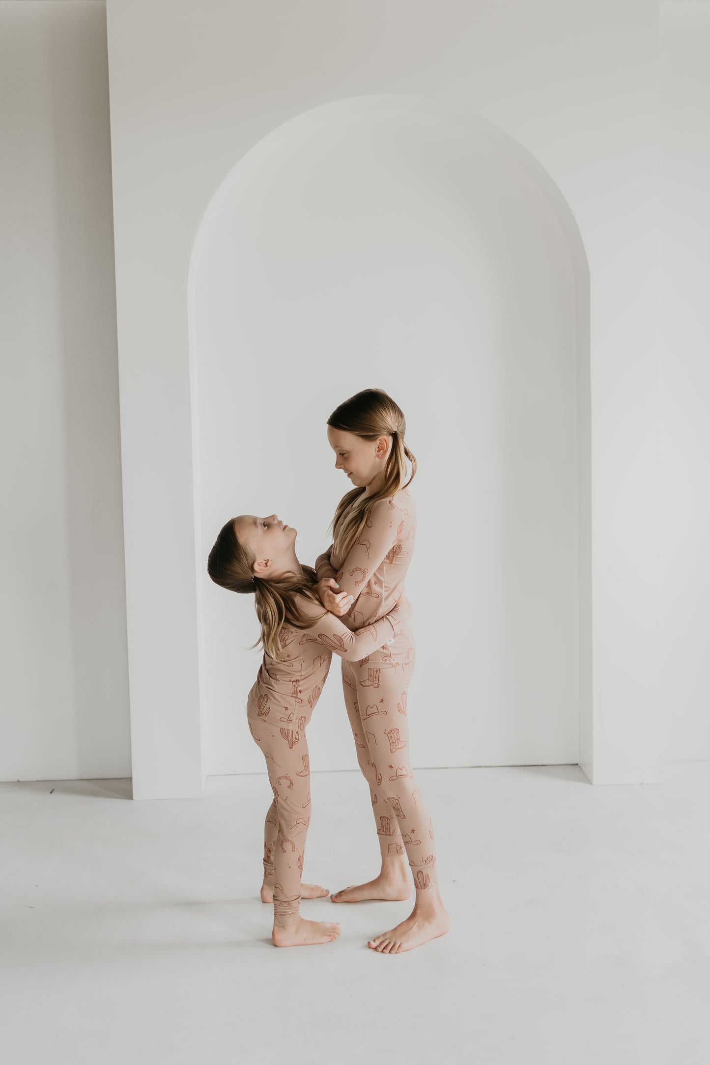 Two children wearing matching beige, hypo-allergenic Bamboo Two Piece Pajamas from the Minty x FF Wild West Desert Dreams collection stand in a white room with an arched backdrop. The older child embraces the younger one, both smiling and looking at each other affectionately.