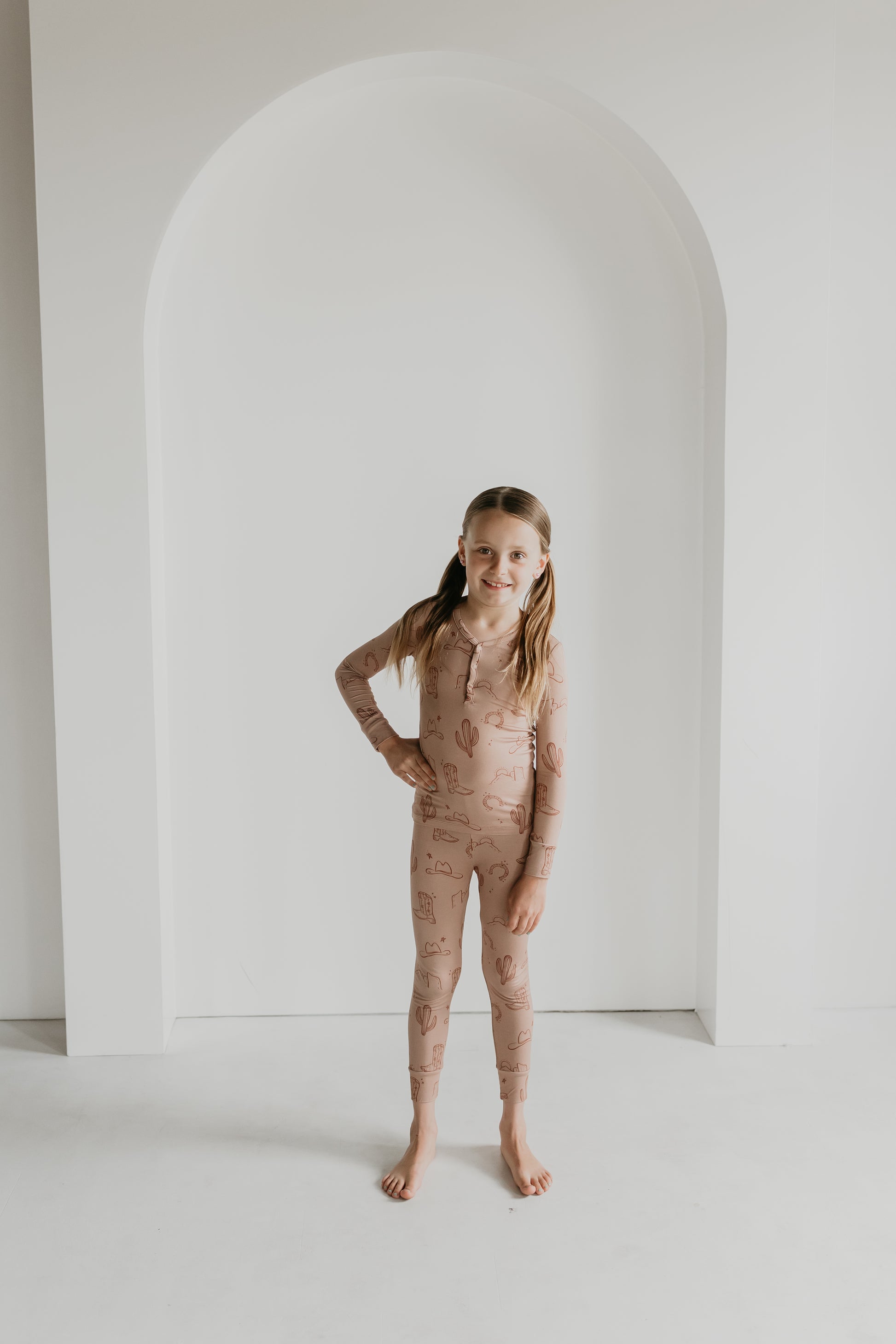 A young girl with long hair stands barefoot in a minimalist setting, wearing the Bamboo Two Piece Pajamas from the Minty x FF Wild West Desert Dreams collection. These light brown pajamas feature simple prints and are hypo-allergenic, promising comfort and style for dreamy nights. She smiles at the camera with one hand on her hip, framed by an arched white wall.