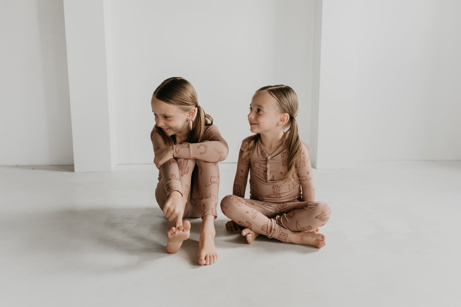 Two young girls sit on the floor against a white wall, both dressed in matching Minty x FF Wild West Desert Dreams Bamboo Two Piece Pajamas with an animal print. The girl on the left sits with her knees bent, resting her arms on them, while the girl on the right sits cross-legged, looking at her sister and smiling.