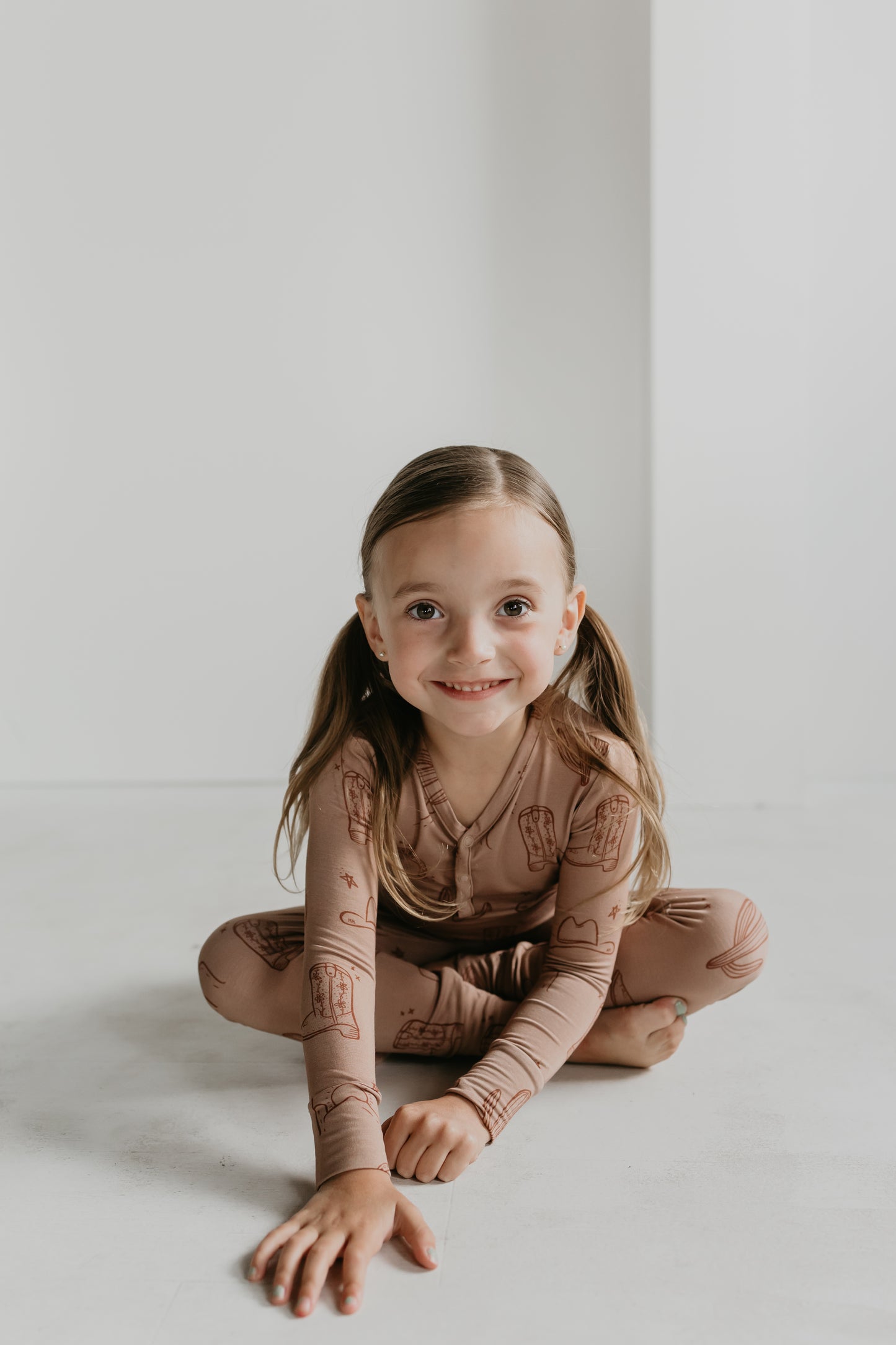 A smiling young girl with long brown hair tied in pigtails sits on the floor with one hand touching the ground. She is wearing the Minty x FF Wild West Desert Dreams Bamboo Two Piece Pajamas, featuring a playful Wild West print. The background is a plain white wall.