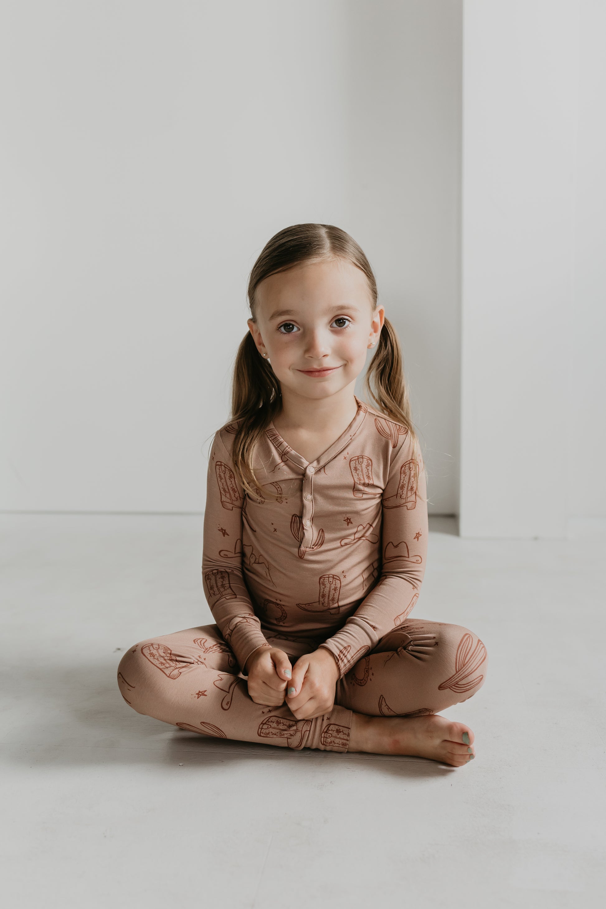 A young girl with light brown hair in pigtails sits cross-legged on the floor, dressed in a matching pink outfit from the Minty x FF Wild West Desert Dreams collection featuring cactus patterns. She smiles gently at the camera. The minimalistic background with light-colored walls and floor complements her serene pose.