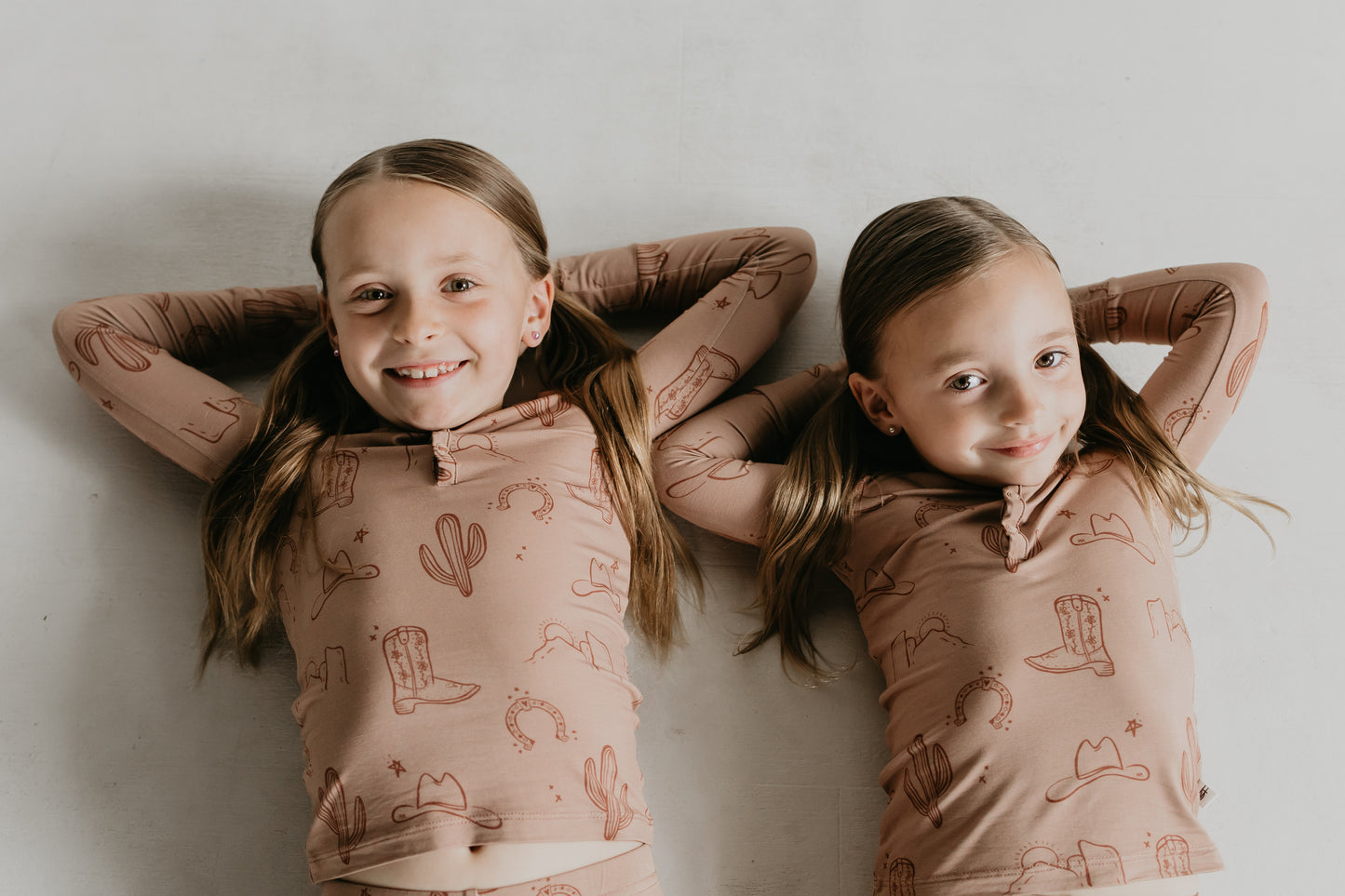 Two young children lie side by side, smiling and resting their hands behind their heads. They wear matching hypo-allergenic Bamboo Two Piece Pajamas | Minty x FF Wild West Desert Dreams prints featuring cacti, cowboy boots, and horseshoes. The background is a plain white surface.