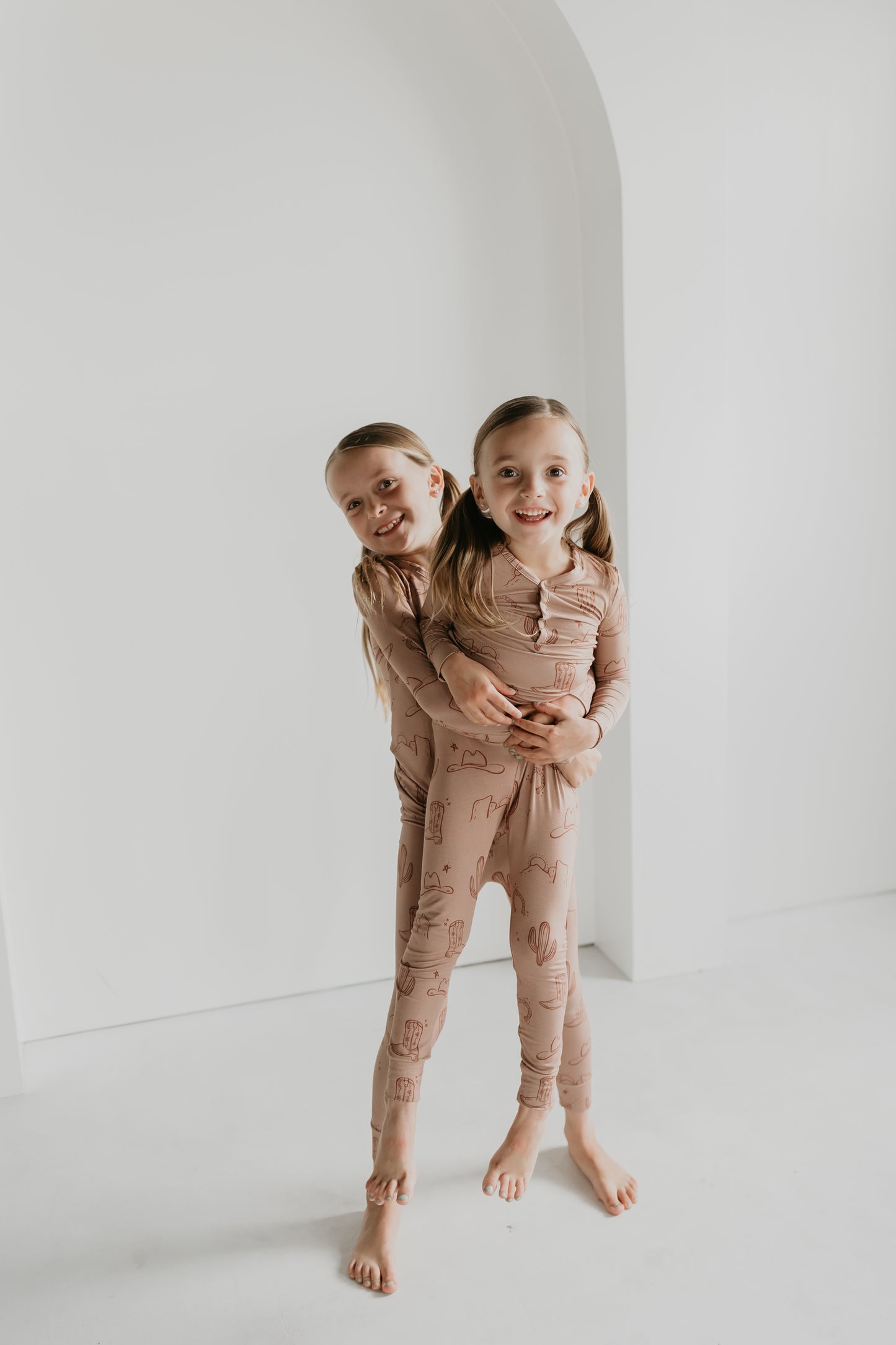 Two smiling young children with long hair are standing barefoot and hugging each other inside a minimalistic white room. They are both wearing matching Bamboo Two Piece Pajamas from Minty x FF Wild West Desert Dreams, featuring a playful print, as they pose playfully for the photo.