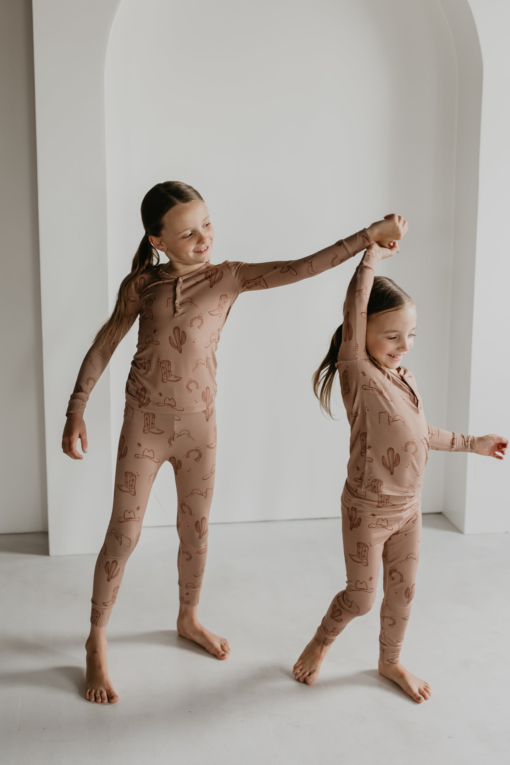 Two children with long hair in braided pigtails stand barefoot in a minimalistic indoor setting, wearing matching brown, hypo-allergenic Bamboo Two Piece Pajamas from the Minty x FF Wild West Desert Dreams collection, adorned with simple cactus motifs. One child, slightly taller, holds the other's raised hand as they smile and dance together, embodying Wild West Desert Dreams.