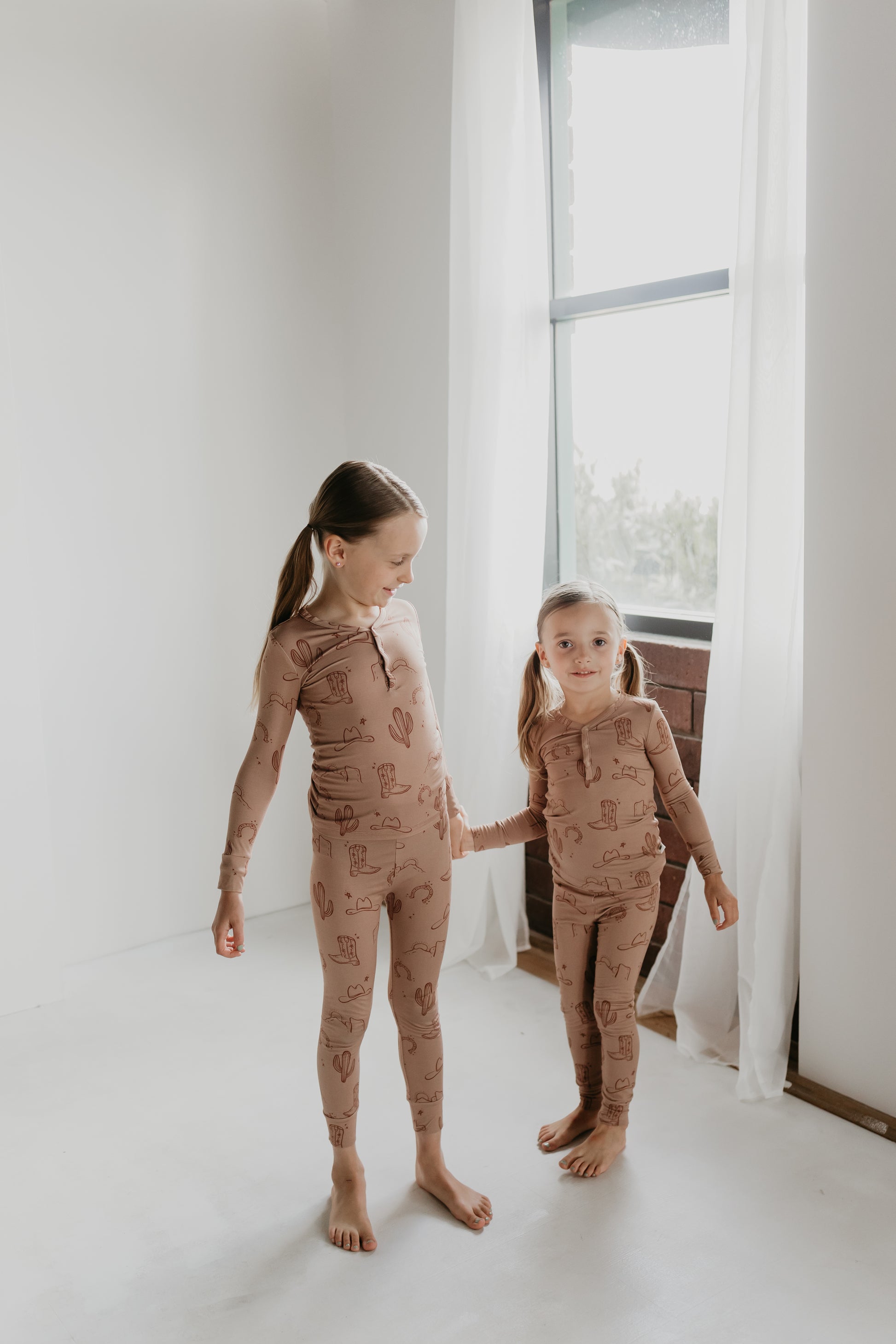 Two young girls stand barefoot in front of a window with white curtains. They are both wearing matching Bamboo Two Piece Pajamas from the Minty x FF Wild West Desert Dreams collection, featuring a playful print design. The older girl is looking down at the younger girl while holding her hand. The room has a minimalistic, serene atmosphere.