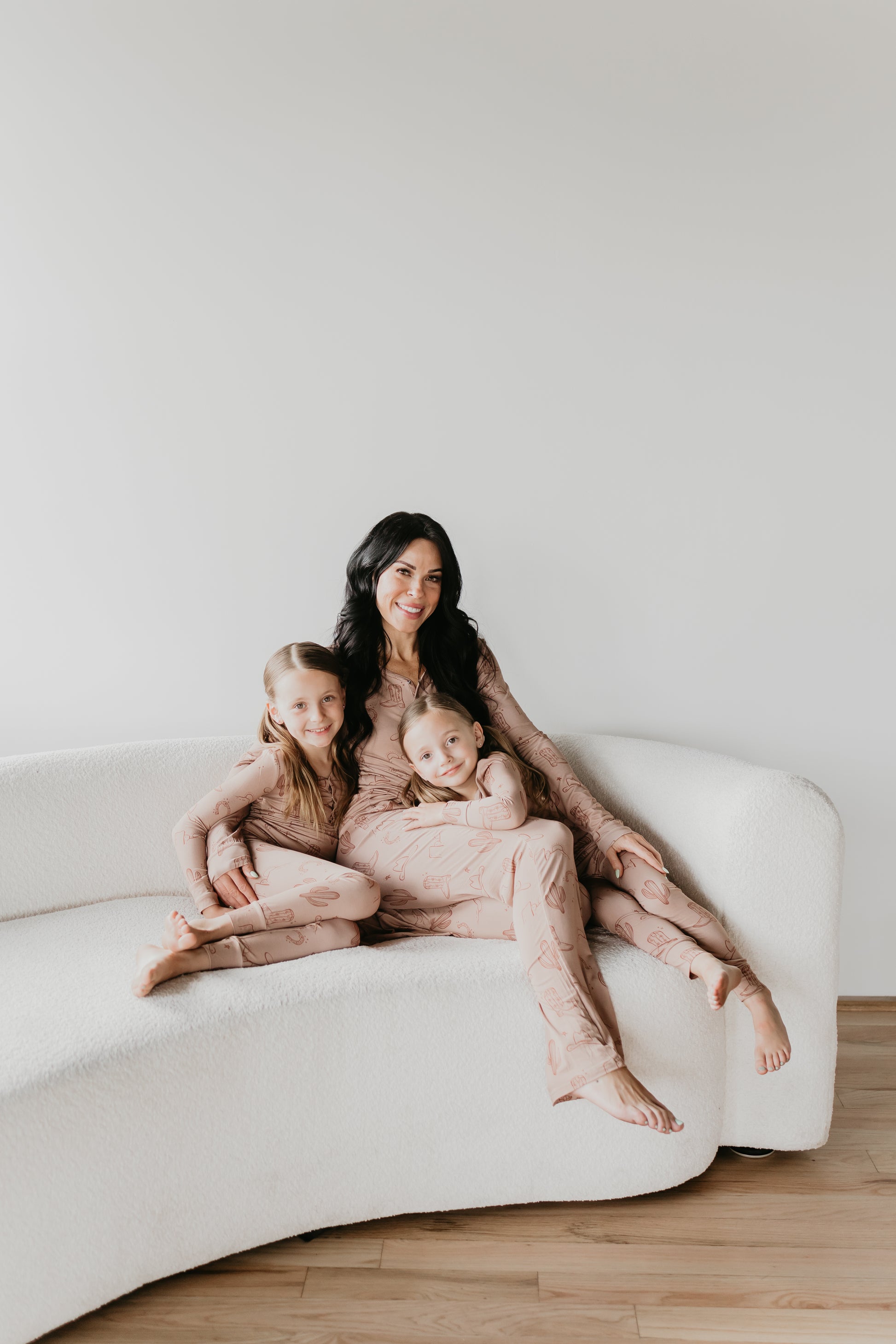 A woman and two children are sitting on a white, curved sofa. They are all wearing matching pajamas from the Minty x FF Wild West Desert Dreams collection, specifically Women's Bamboo Pajamas in a breathable fabric with abstract patterns. The woman is smiling at the camera while the children lean against her, appearing happy and relaxed. The backdrop is a clean, white wall.