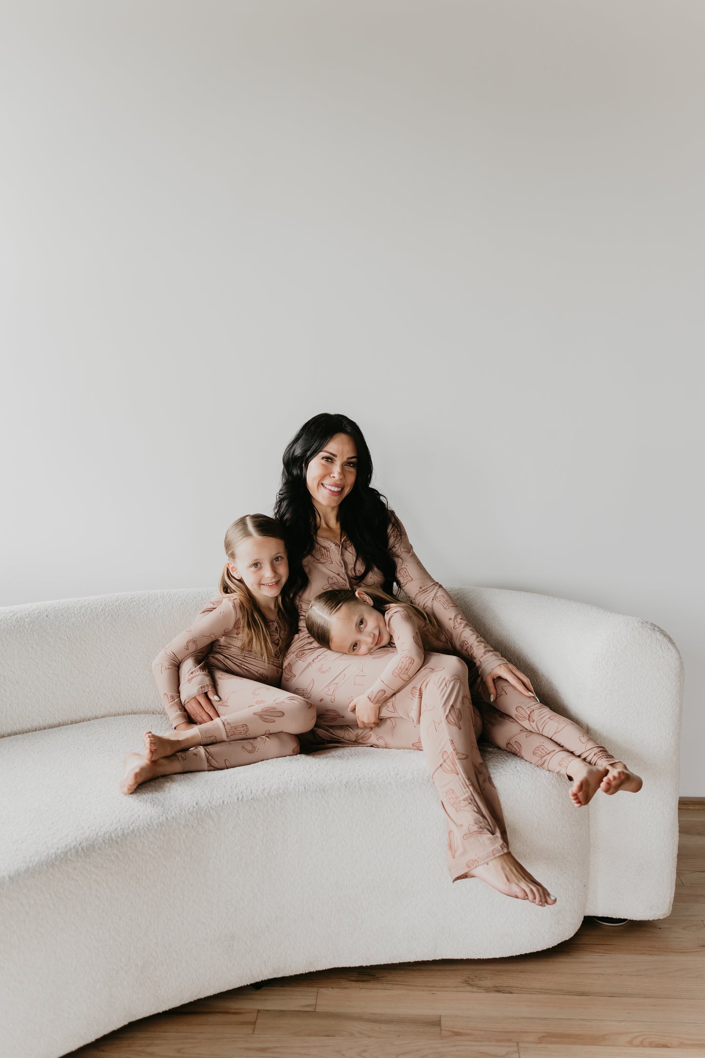 A woman with long dark hair sits on a curved white sofa with two young girls on either side of her, all wearing matching Minty x FF Wild West Desert Dreams Bamboo Two Piece Pajamas. The girls are leaning affectionately towards the smiling woman, framed by a plain white wall.