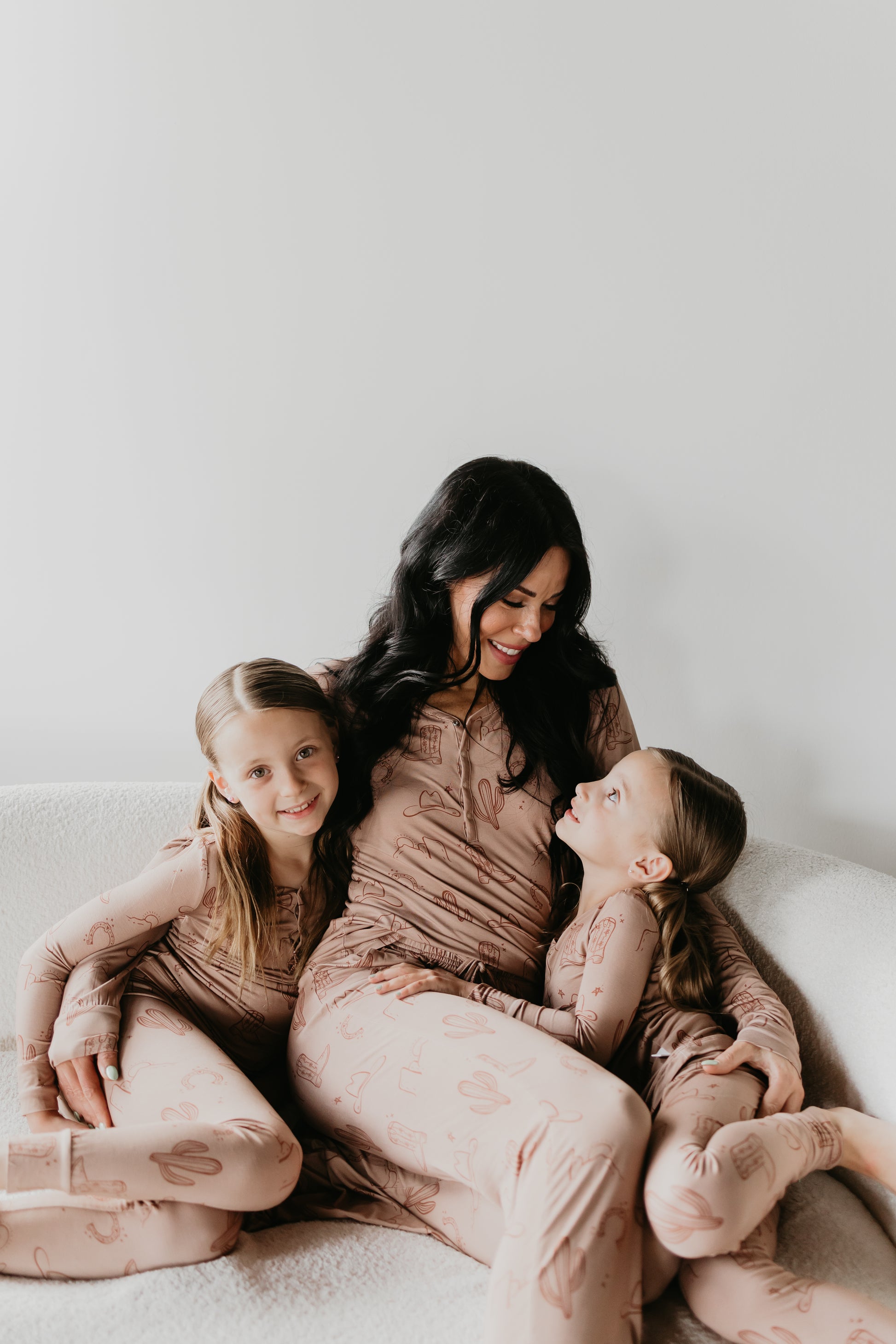 A woman with long dark hair sits on a white couch, smiling at two young girls. All three are wearing matching hypo-allergenic Bamboo Two Piece Pajamas from Minty x FF Wild West Desert Dreams, adorned with a faint pattern. The girls, sitting on either side of the woman, are looking affectionately towards her.