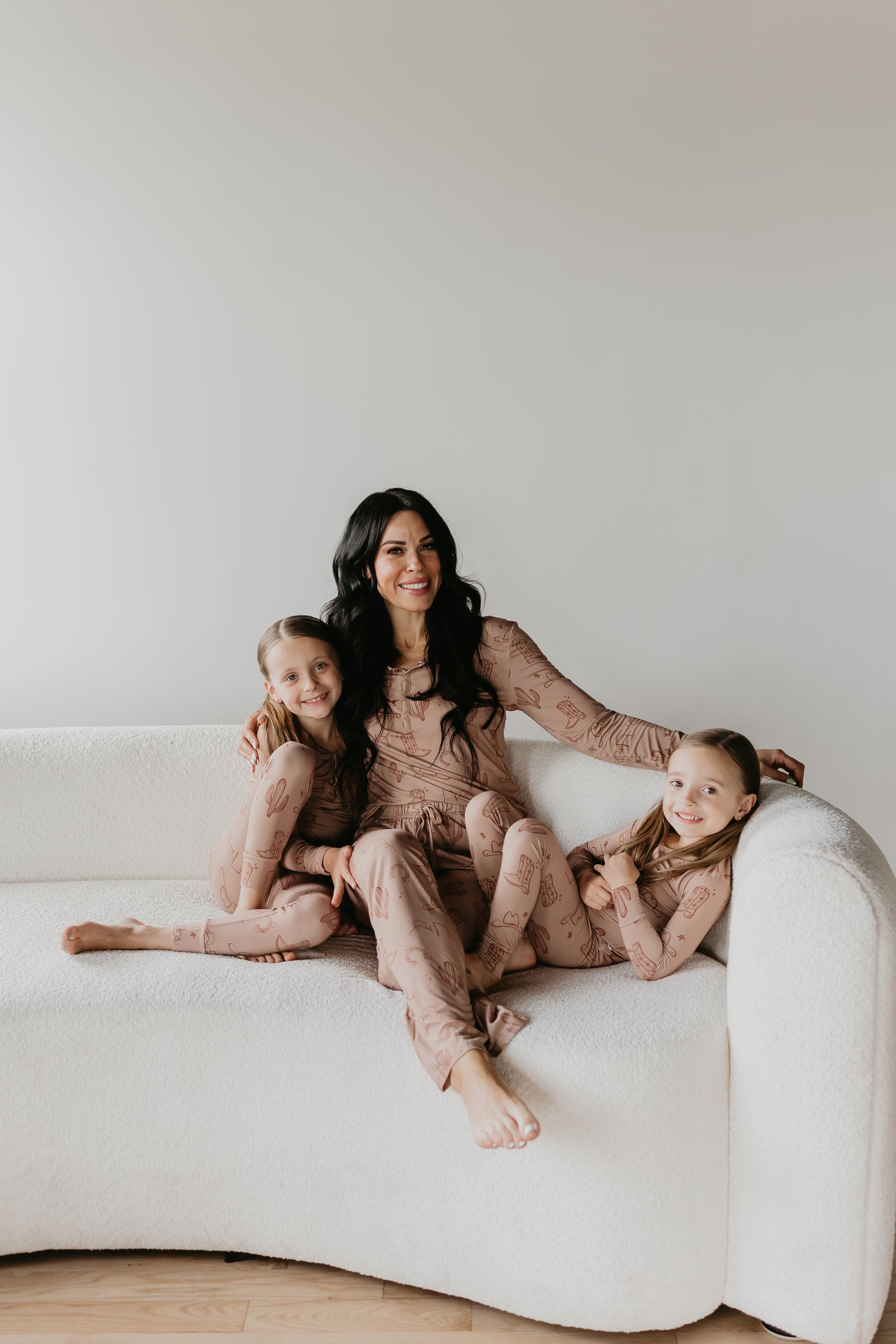 A woman with long dark hair sits on a white sofa, smiling and embracing two young girls with matching blonde hair. All three are wearing light brown Women's Bamboo Pajamas from the Minty x FF Wild West Desert Dreams collection, featuring a breathable fabric with a doodle pattern. The girls are seated on either side of the woman, also smiling and appearing cheerful.