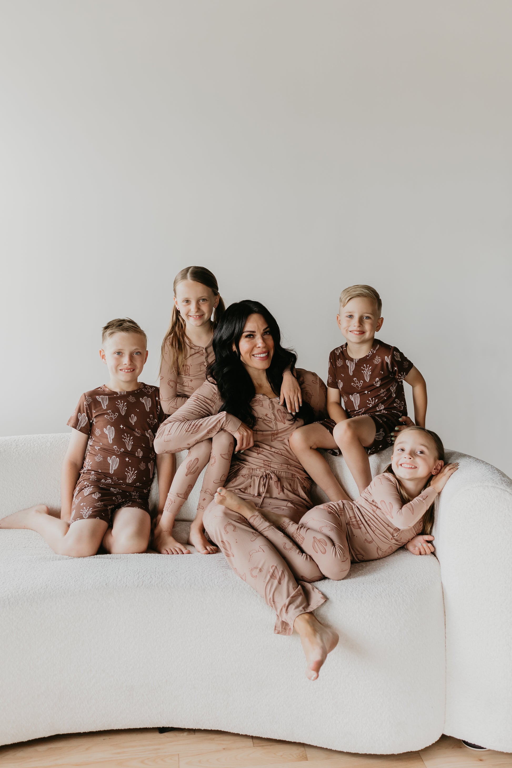 A woman with dark hair sits on a white couch, surrounded by four children. Two boys and two girls, all wearing matching outfits from the Minty x FF Wild West Desert Dreams collection, smile and pose with her. Their Bamboo Two Piece Pajamas add a touch of coordinated charm to the minimalist and cozy setting against the plain white background.