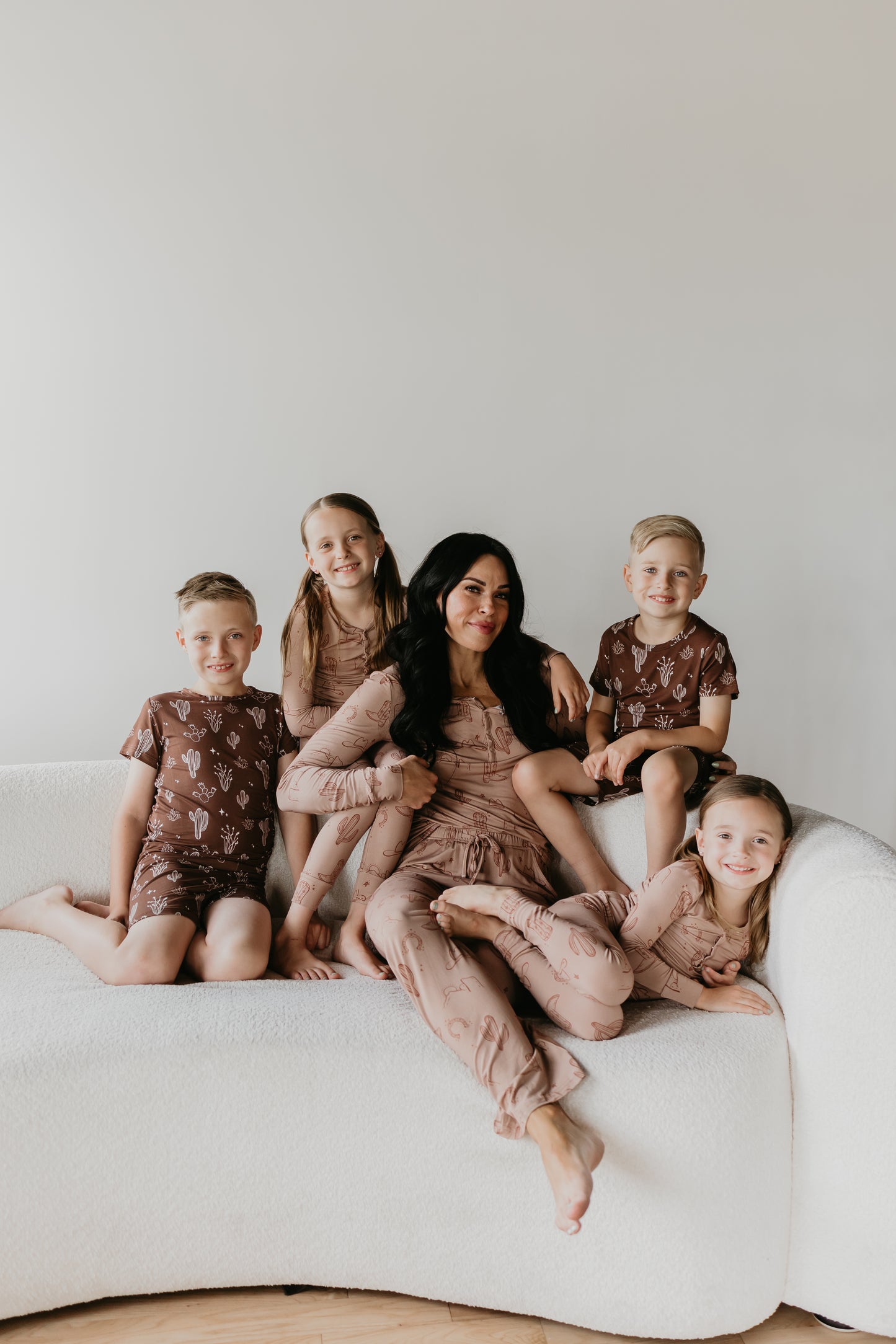 A woman sits on a white couch with four smiling children around her, all wearing matching breathable pajamas adorned with cactus patterns. The soft Women’s Bamboo Pajamas from Minty x FF Wild West Desert Dreams ensure everyone is comfortable, and the hypo-allergenic fabric adds an extra layer of care. The background is plain white.