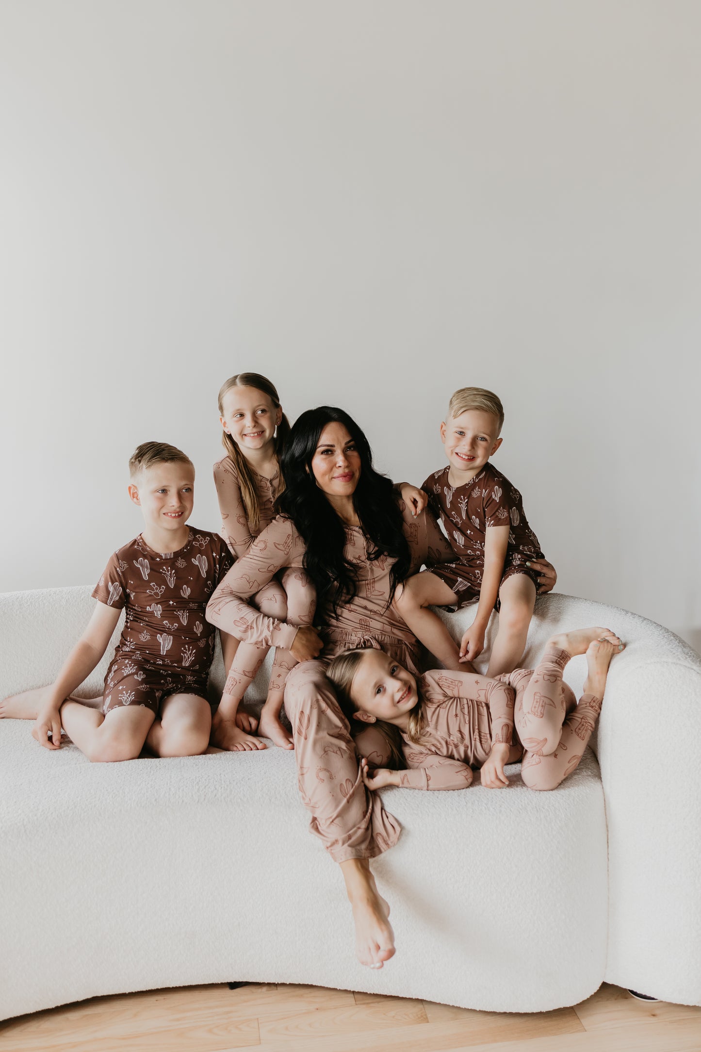 A woman with long, dark hair sits on a white sofa with four children around her, all wearing matching earth-toned Women's Bamboo Pajamas from the Minty x ff Wild West Desert Dreams collection. The children are smiling and striking playful poses next to the woman. The background is simple and neutral.