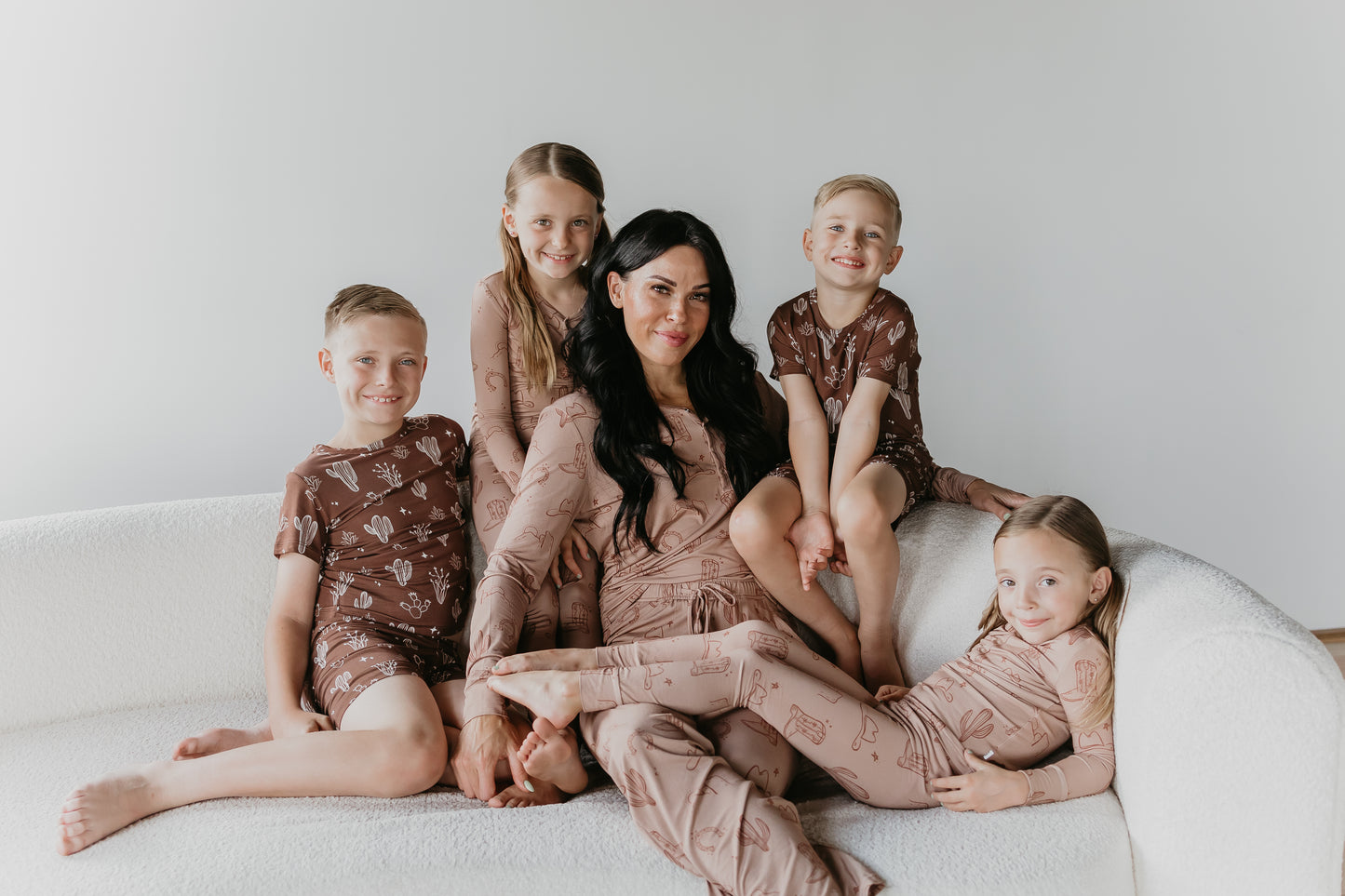 A woman sits on a white sofa with four children, all wearing matching brown outfits made from breathable fabric with subtle patterns. The woman, dressed in Women's Bamboo Pajamas from Minty x FF Wild West Desert Dreams, is in the center, surrounded by two children on each side. They are all smiling and looking at the camera against a plain white background.