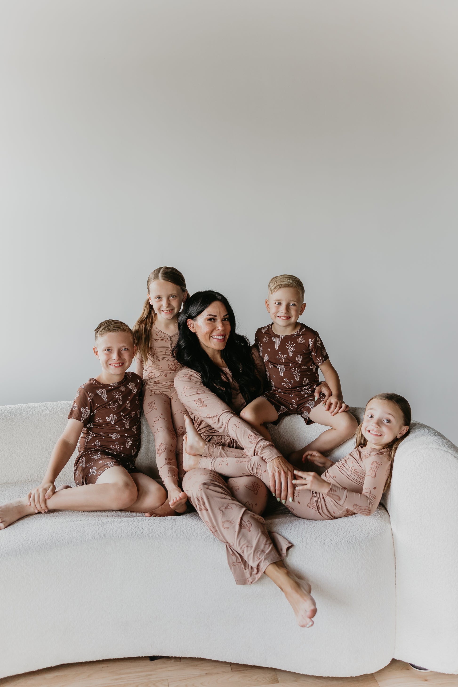 A woman with long dark hair sits on a white couch with four smiling children dressed in matching Minty x ff Wild West Women's Bamboo Pajamas. The group appears happy and relaxed against a plain light-colored background.