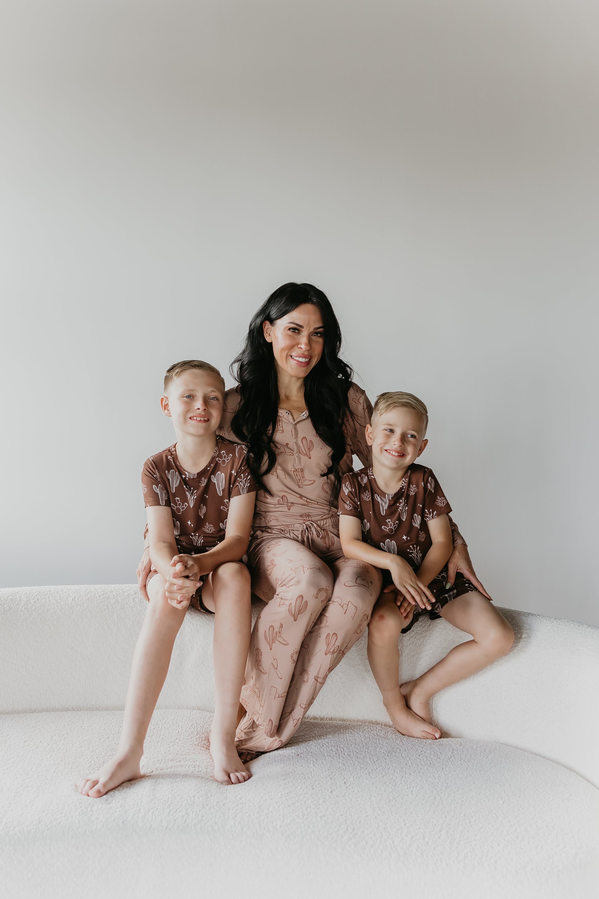 A woman in breathable fabric sits on a white couch with two young boys, all smiling. She wears Women's Bamboo Pajamas by Minty x FF Wild West Desert Dreams, featuring a subtle print, while the boys are dressed in matching hypo-allergenic clothing with a similar design. The background is plain white, creating a clean and simple setting.