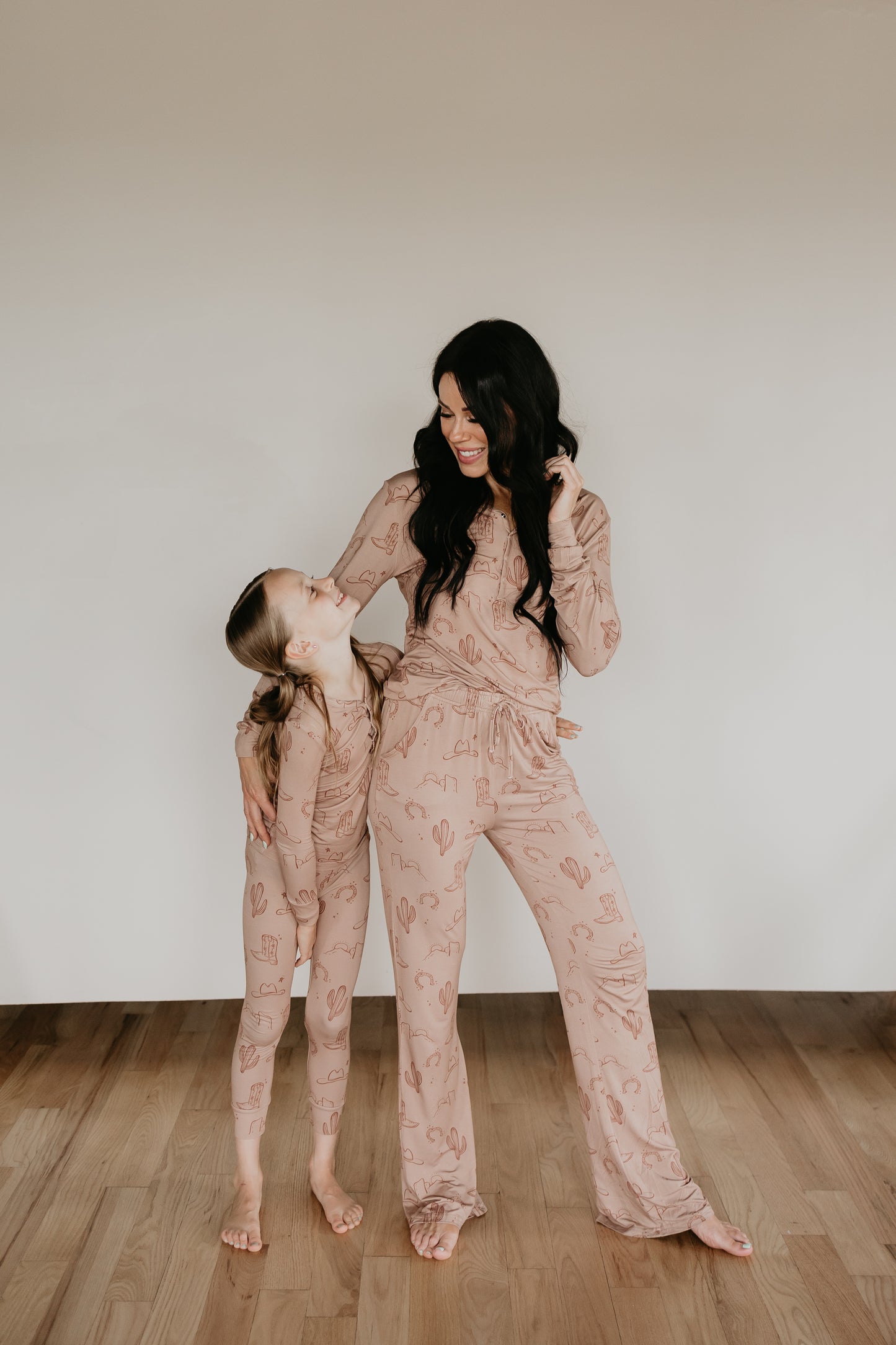 A woman and a young girl stand on a wooden floor, smiling and looking at each other. Both are barefoot and wearing matching light-colored Women's Bamboo Pajamas from the Minty x FF Wild West Desert Dreams collection, featuring cactus patterns and made from breathable fabric. The woman has long dark hair, while the girl has her blonde hair in a braid.