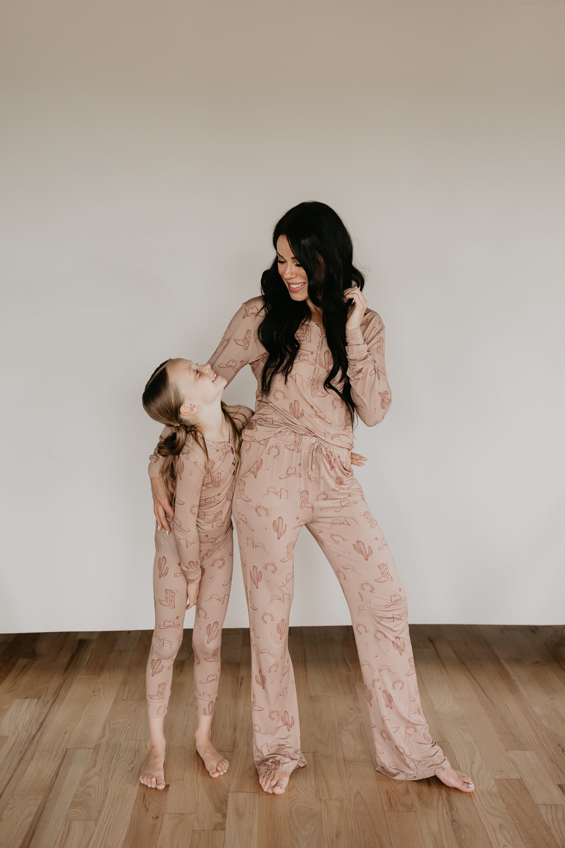 A woman and a young girl stand on a wooden floor, smiling and looking at each other. Both are barefoot and wearing matching light-colored Women's Bamboo Pajamas from the Minty x FF Wild West Desert Dreams collection, featuring cactus patterns and made from breathable fabric. The woman has long dark hair, while the girl has her blonde hair in a braid.