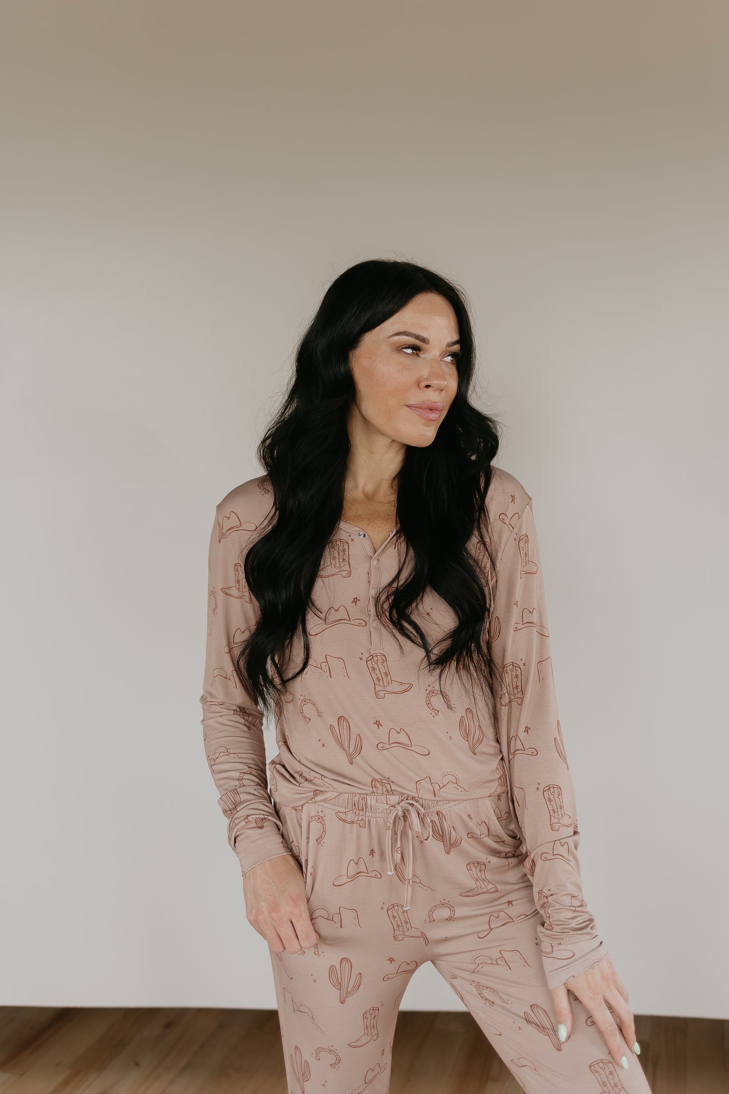 A woman with long, dark hair stands against a plain background wearing a matching Women's Bamboo Pajamas set from Minty x FF Wild West Desert Dreams. The light brown set features a cactus and desert-themed print and is made from breathable fabric. She has her left hand in her pocket and is looking slightly to her right with a relaxed expression.