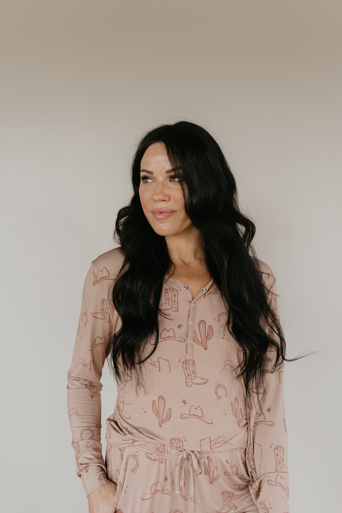 A person with long, wavy dark hair stands against a plain background, wearing the Women’s Bamboo Pajamas from Minty x FF Wild West Desert Dreams in light brown. The outfit features a southwestern print with cacti, cowboy hats, and boots, skillfully crafted from breathable fabric. They have their hands in their pockets and are looking slightly to the left with a soft smile.