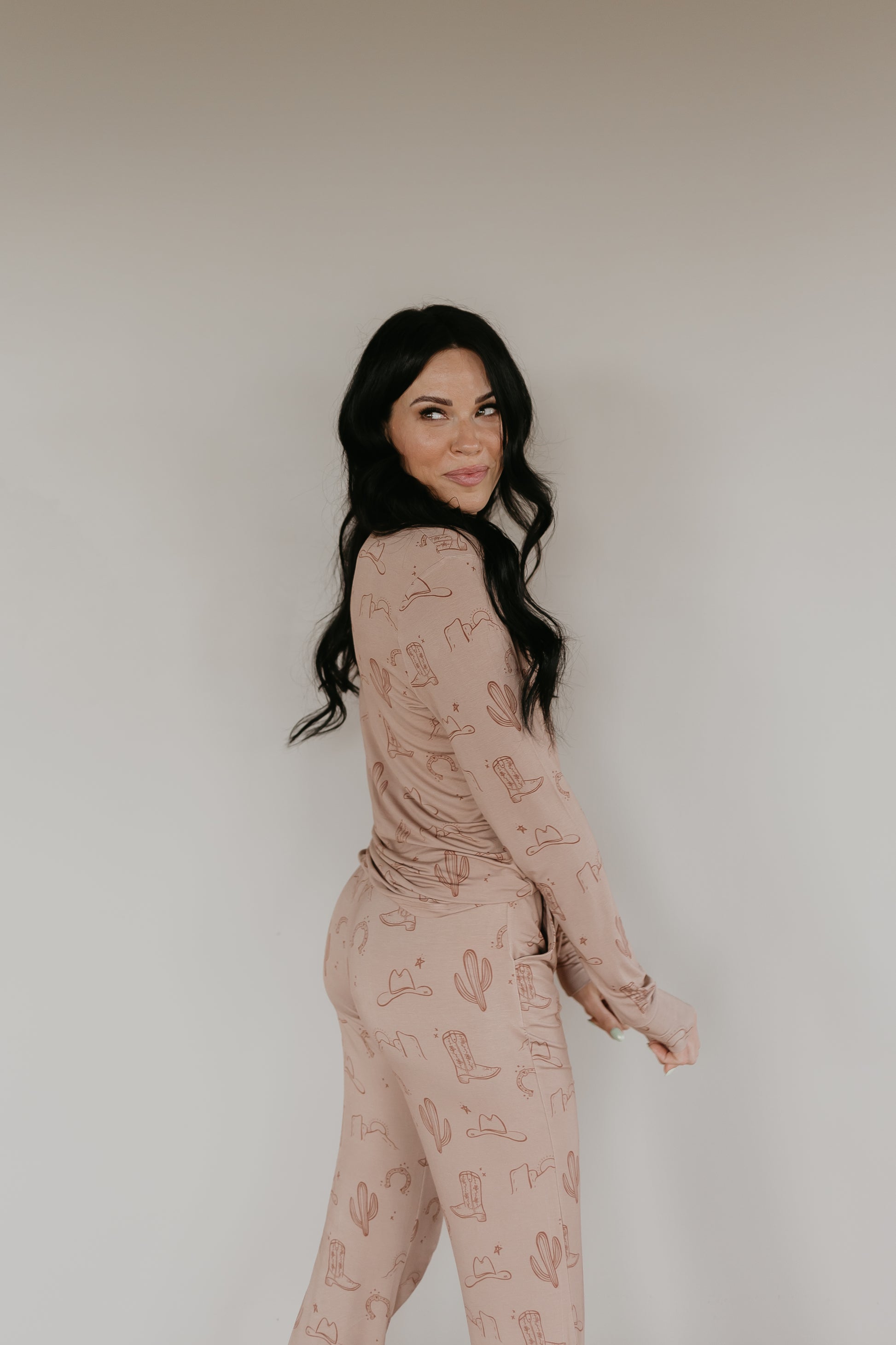 A woman with long, dark hair poses against a plain background, wearing a matching beige pajama set adorned with a cactus pattern. Sporting the Women's Bamboo Pajamas from Minty x FF Wild West Desert Dreams' Minty x ff Wild West collection, the breathable fabric offers her comfort as she smiles and looks over her shoulder.